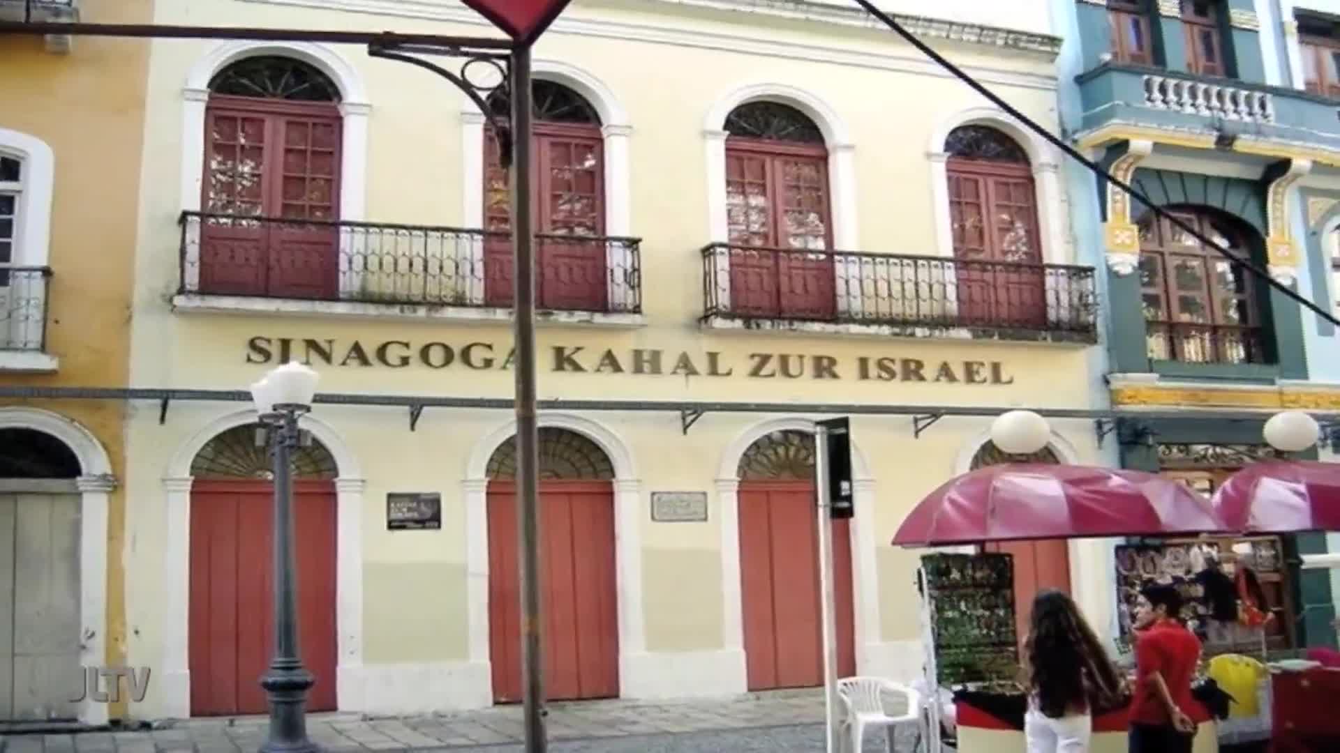 A young woman in white pants and a woman in a red shirt stand near a market stall in front of the Sinagoga Kahal Zur Israel. The building's facade features arched doorways and balconies with ornate railings.