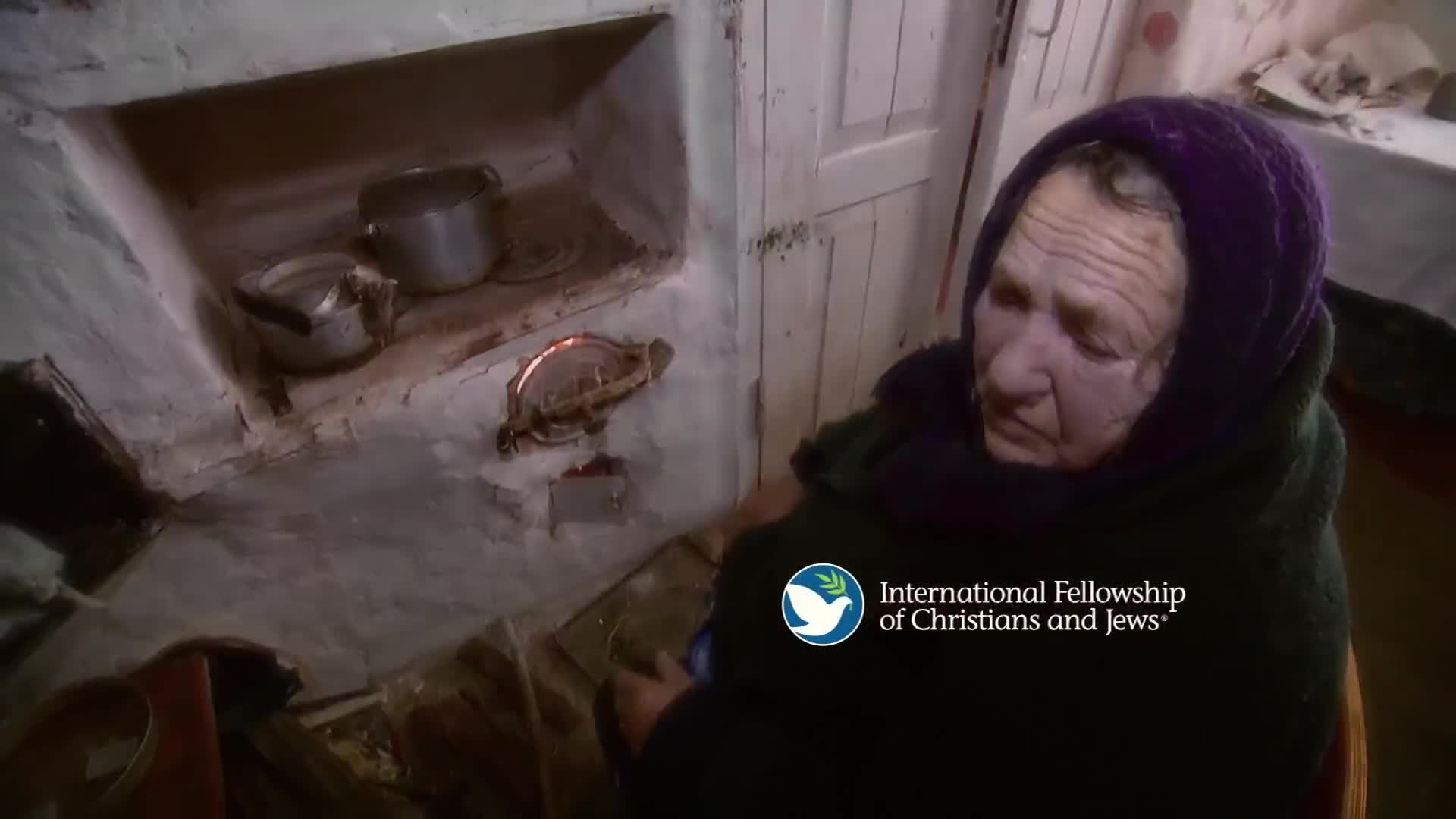 An elderly woman wrapped in a dark shawl sits near a rustic stove. Flames flicker within the stove's opening, casting a warm glow.