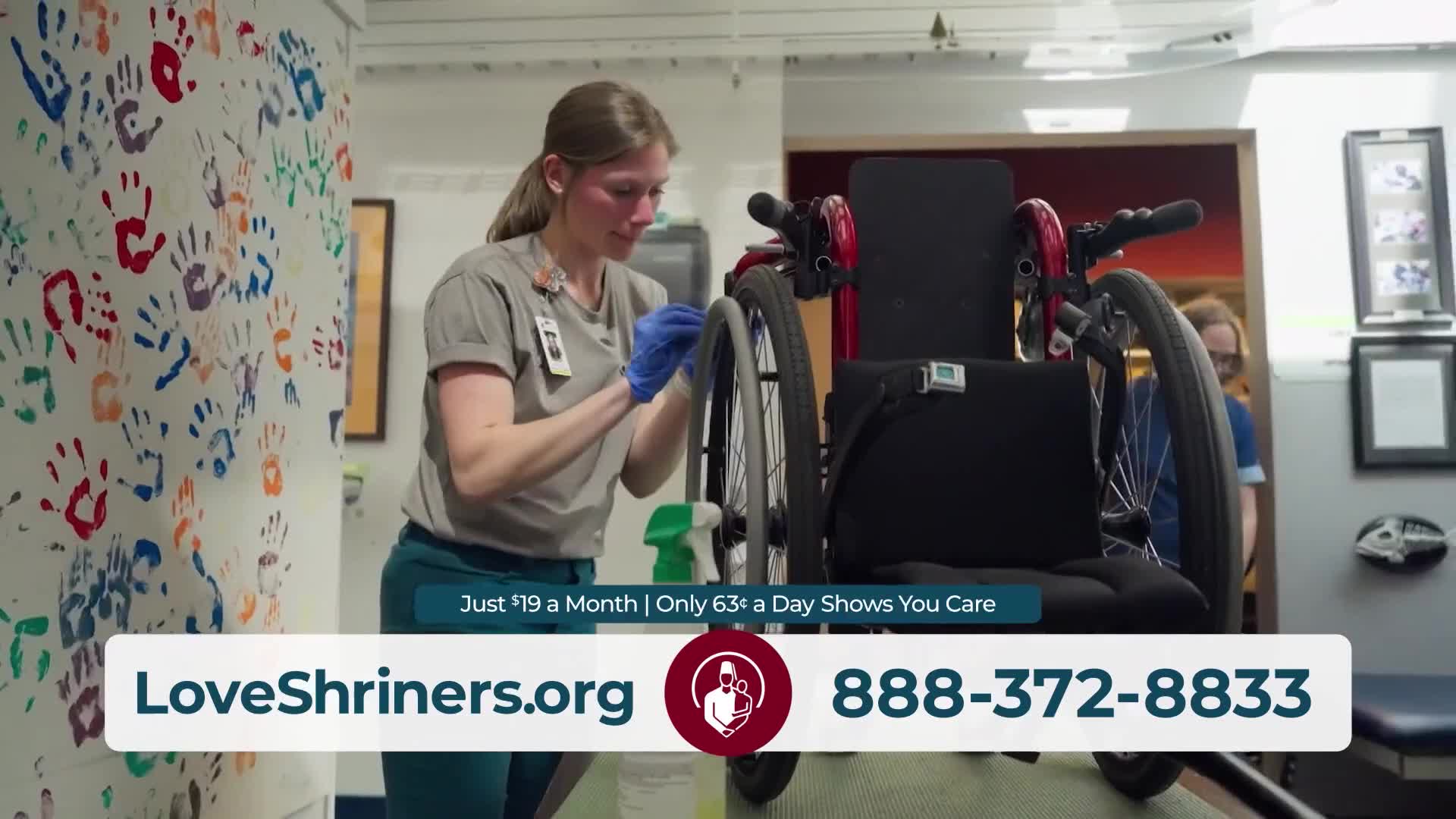 A woman in blue gloves wipes down a wheelchair with a spray bottle. Colorful handprints cover the wall to her left.