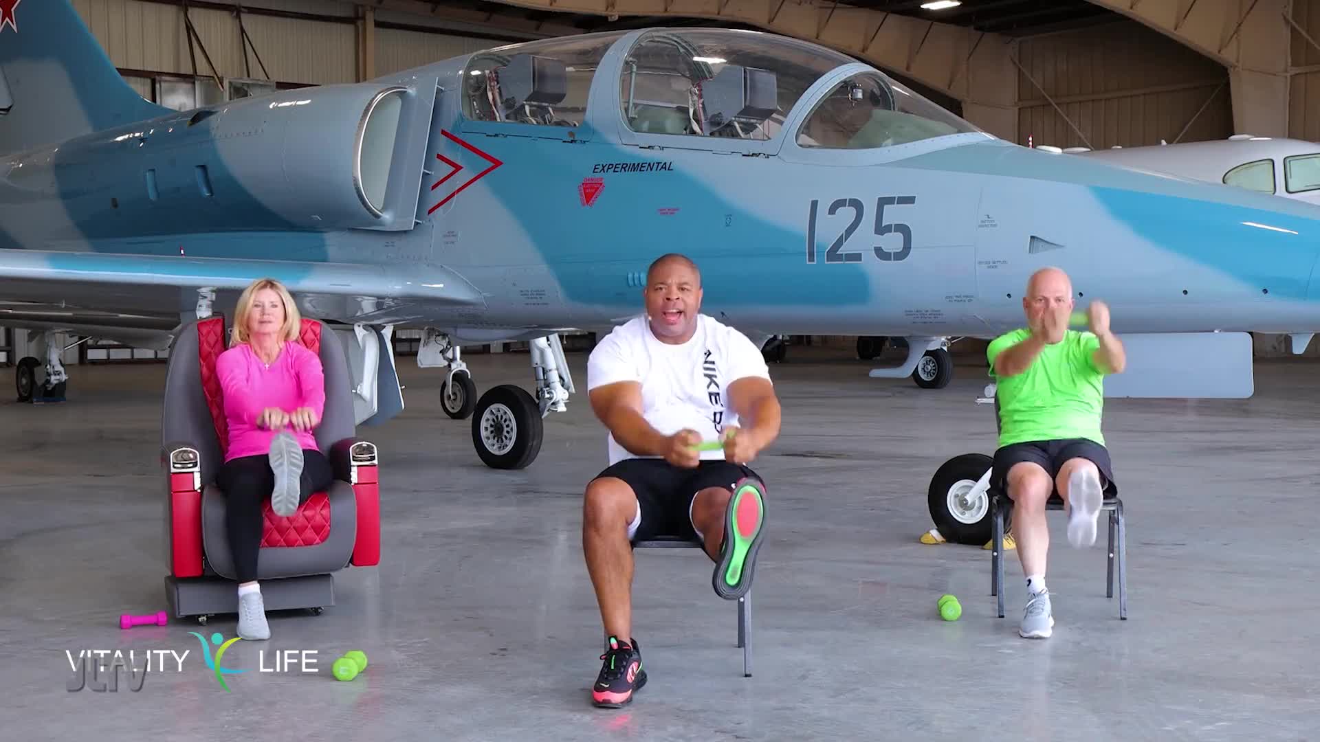 Three people are exercising in chairs in front of a jet plane. The woman on the left is holding her legs out, while the two men in the center and right are pumping their arms and lifting their legs.