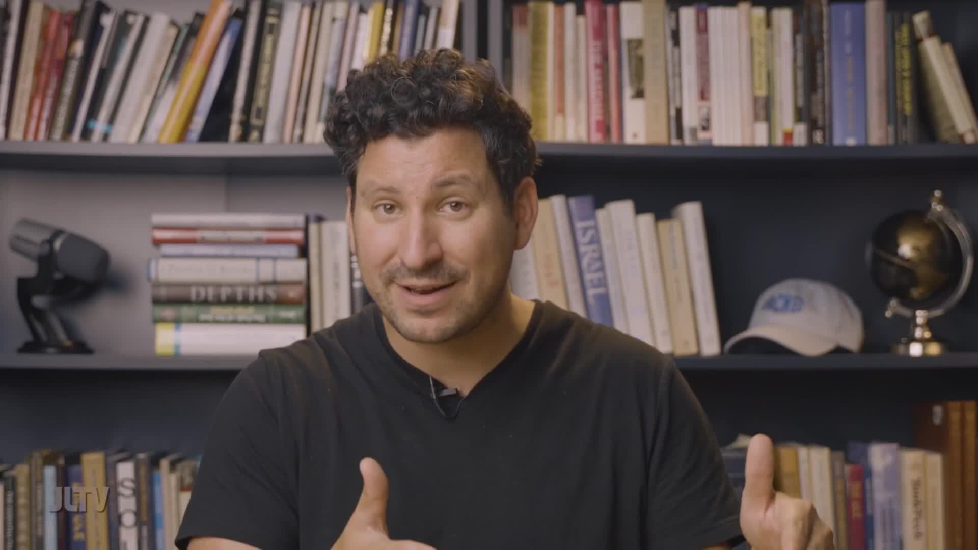 A man speaks directly to the camera, his hands gesturing as he talks. Behind him, bookshelves overflow with volumes, some stacked horizontally.
