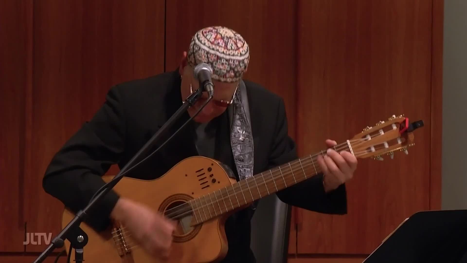 A man in a black jacket and a patterned kippah plays an acoustic guitar, his fingers moving across the fretboard. The JLTV logo is visible in the lower left corner of the screen.