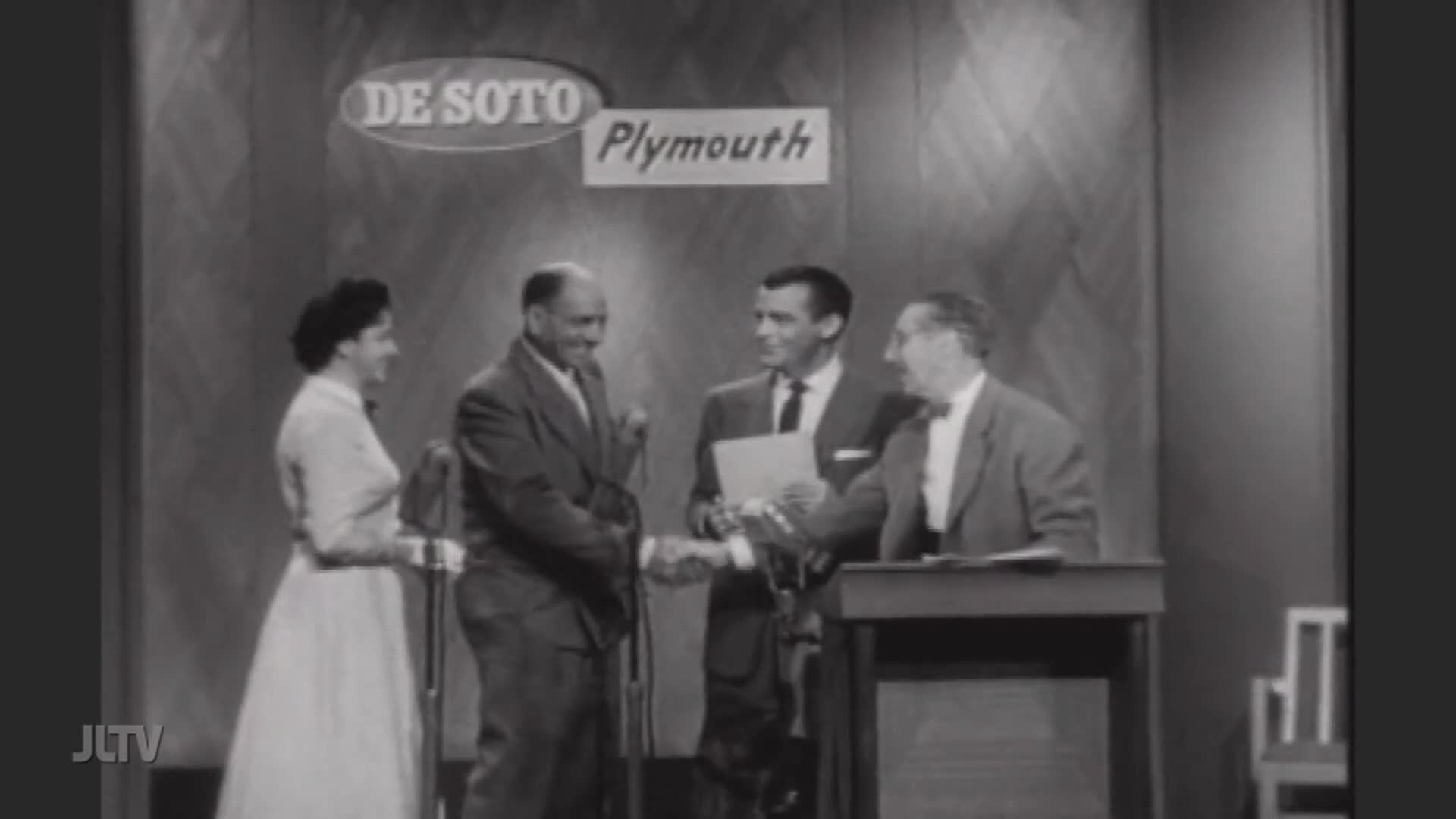 A man in a suit shakes hands with another man, a paper held between them. A woman in a dress stands to the side, smiling.