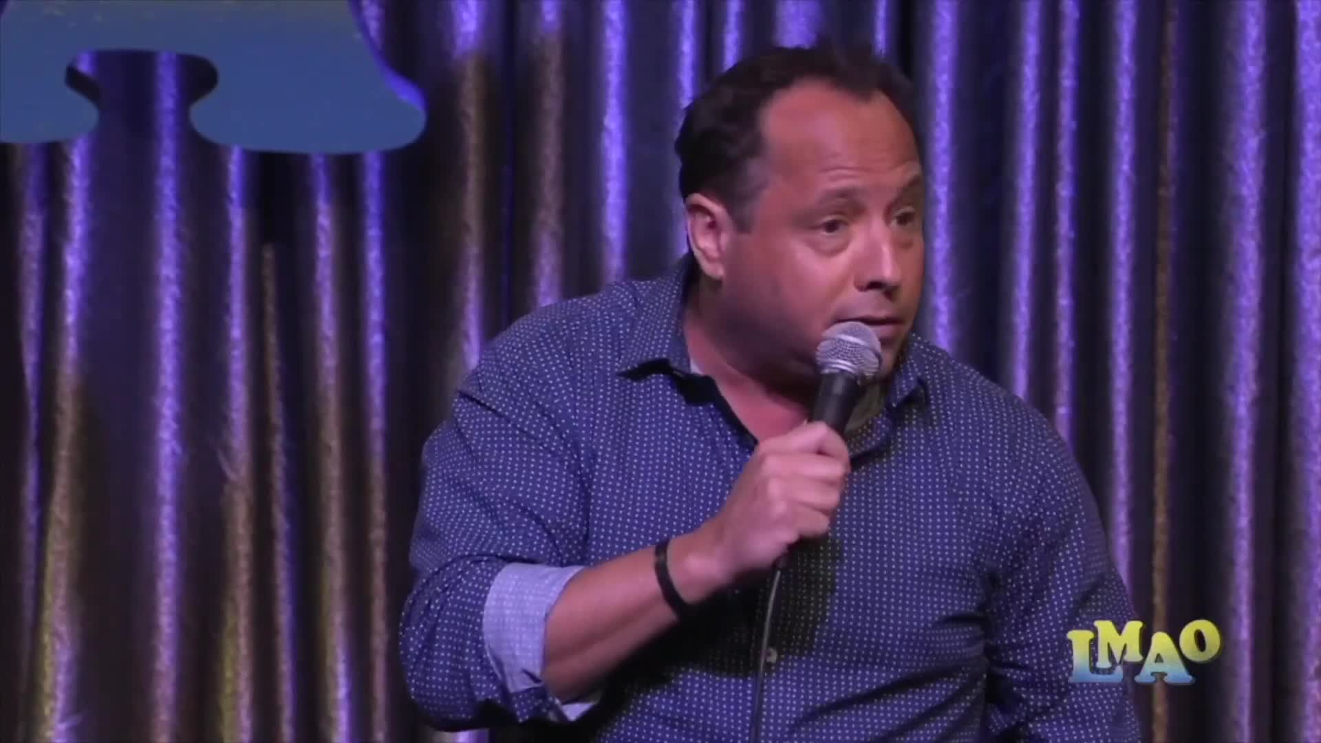 A man holds a microphone, his expression animated as he speaks into it. He is on a stage with a draped purple backdrop.