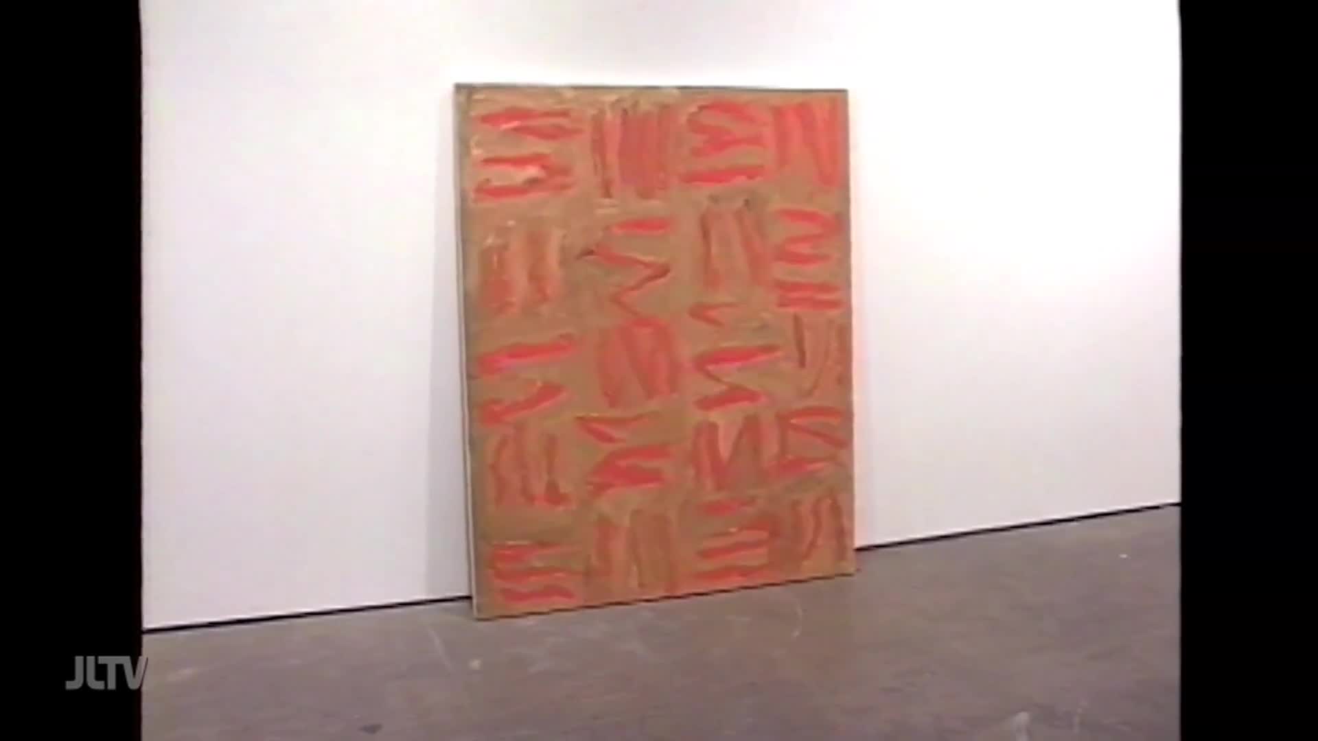 A large, rectangular painting with a tan background and bold red strokes stands leaning against a white wall. The red marks are applied in a grid-like pattern, some appearing as short, thick lines, others as more fluid zigzags.