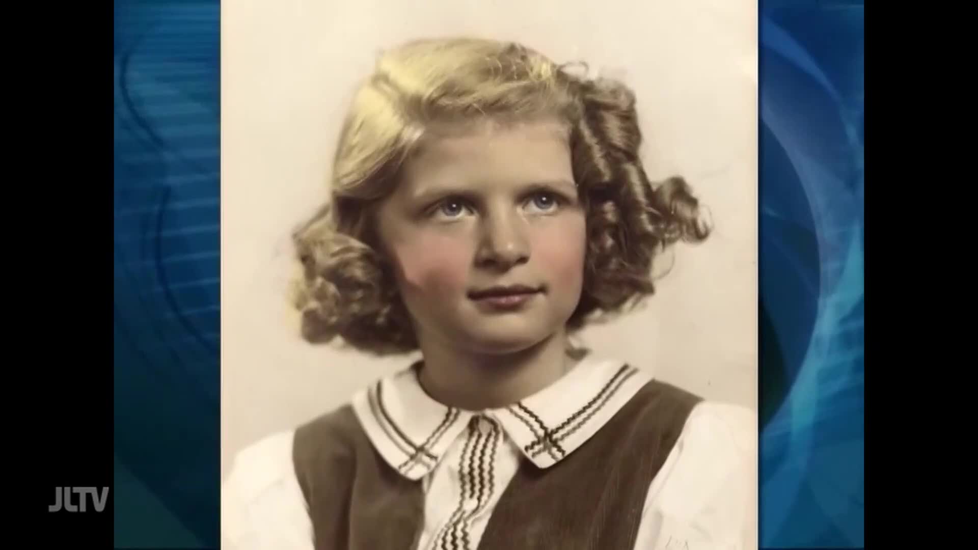 A young girl with blonde curls stares directly ahead. Her cheeks are flushed pink, and she wears a white blouse with a dark pinafore.