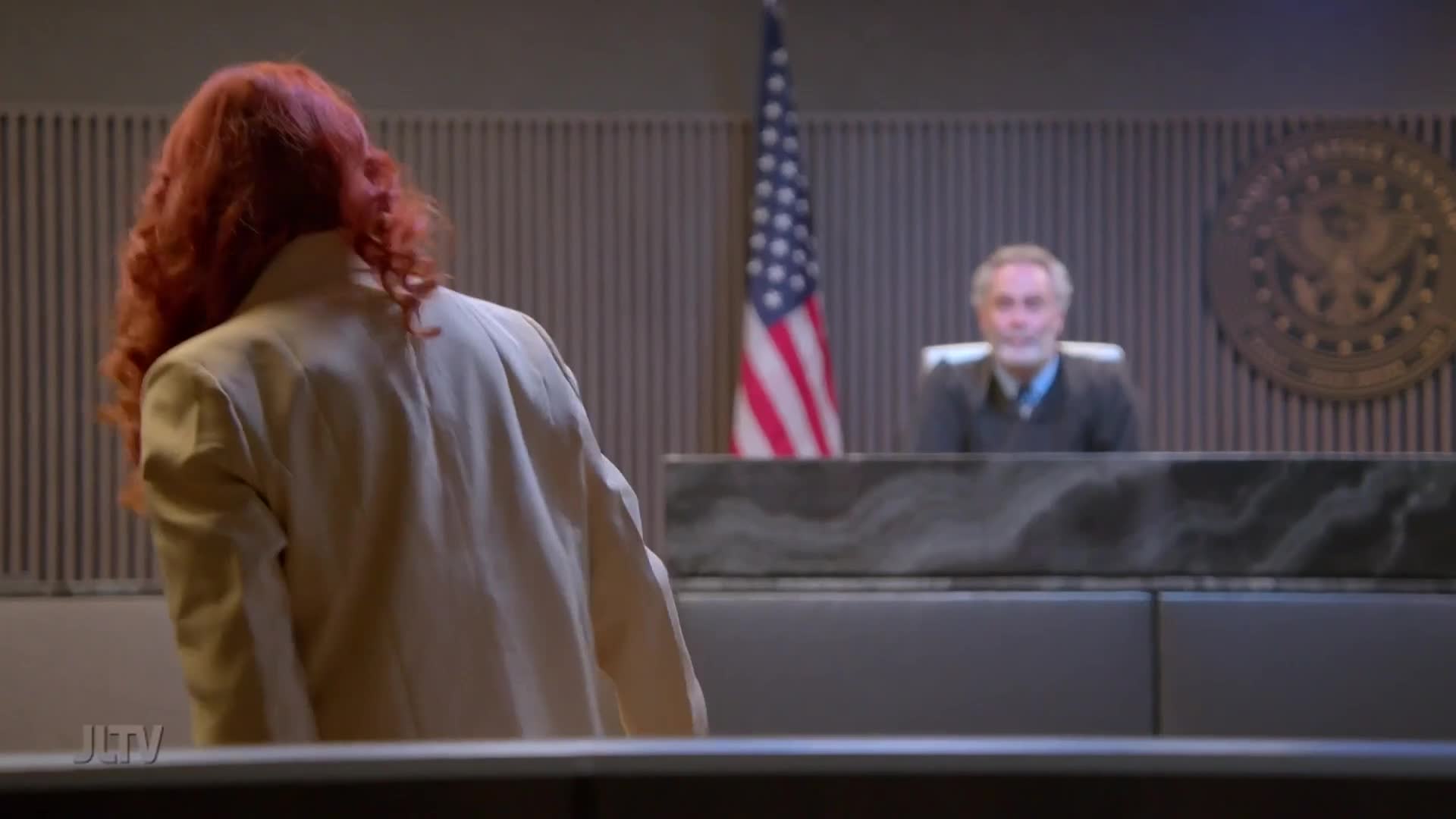 A woman with bright red hair, wearing a tan jacket, turns away from the camera in a courtroom. Behind her, an American flag stands beside a judge seated at his bench.