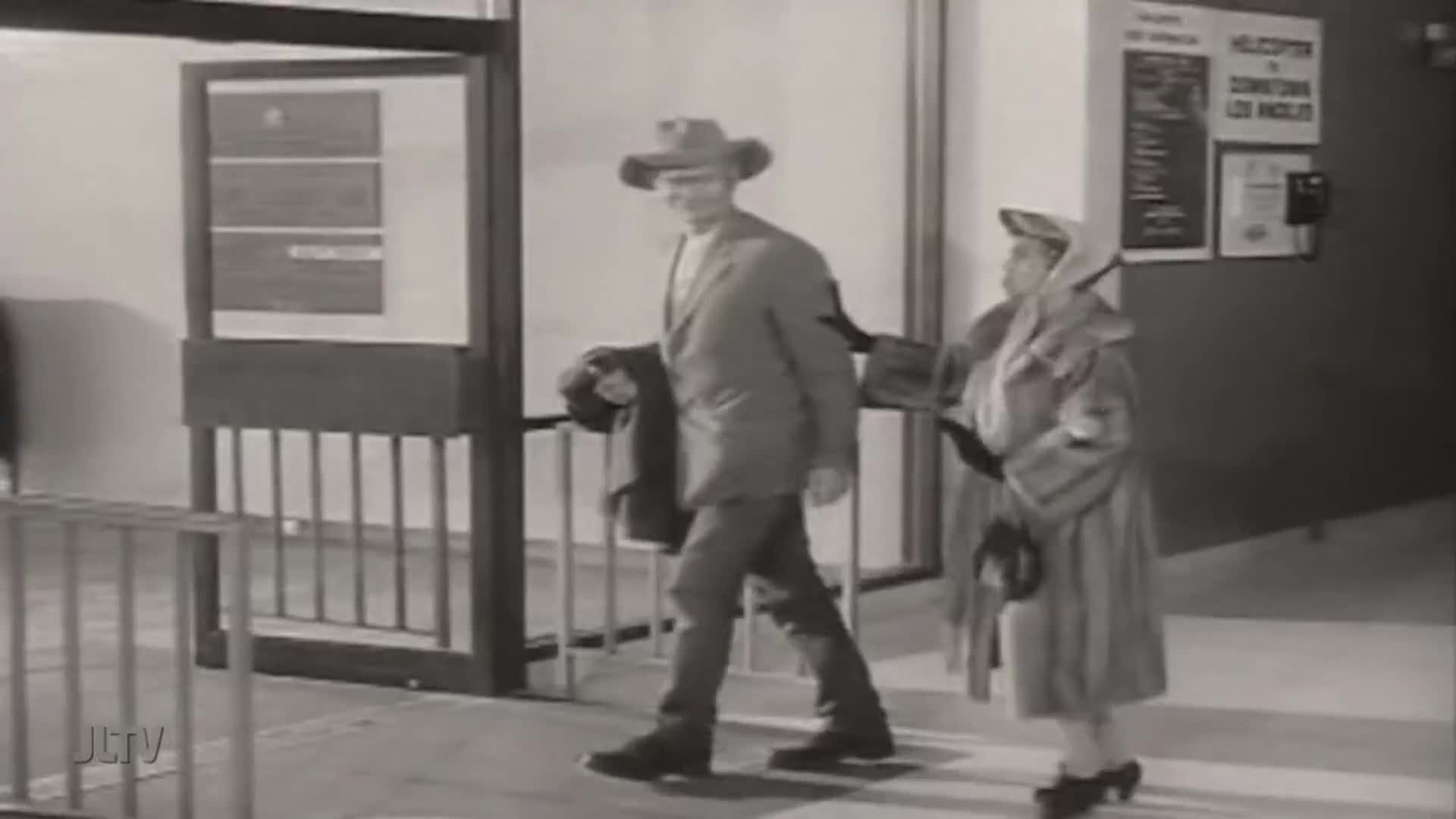 A man in a cowboy hat and suit jacket walks by, a woman in a fur coat following close behind. A sign for "Helicopter to Downtown Los Angeles" hangs on the wall.