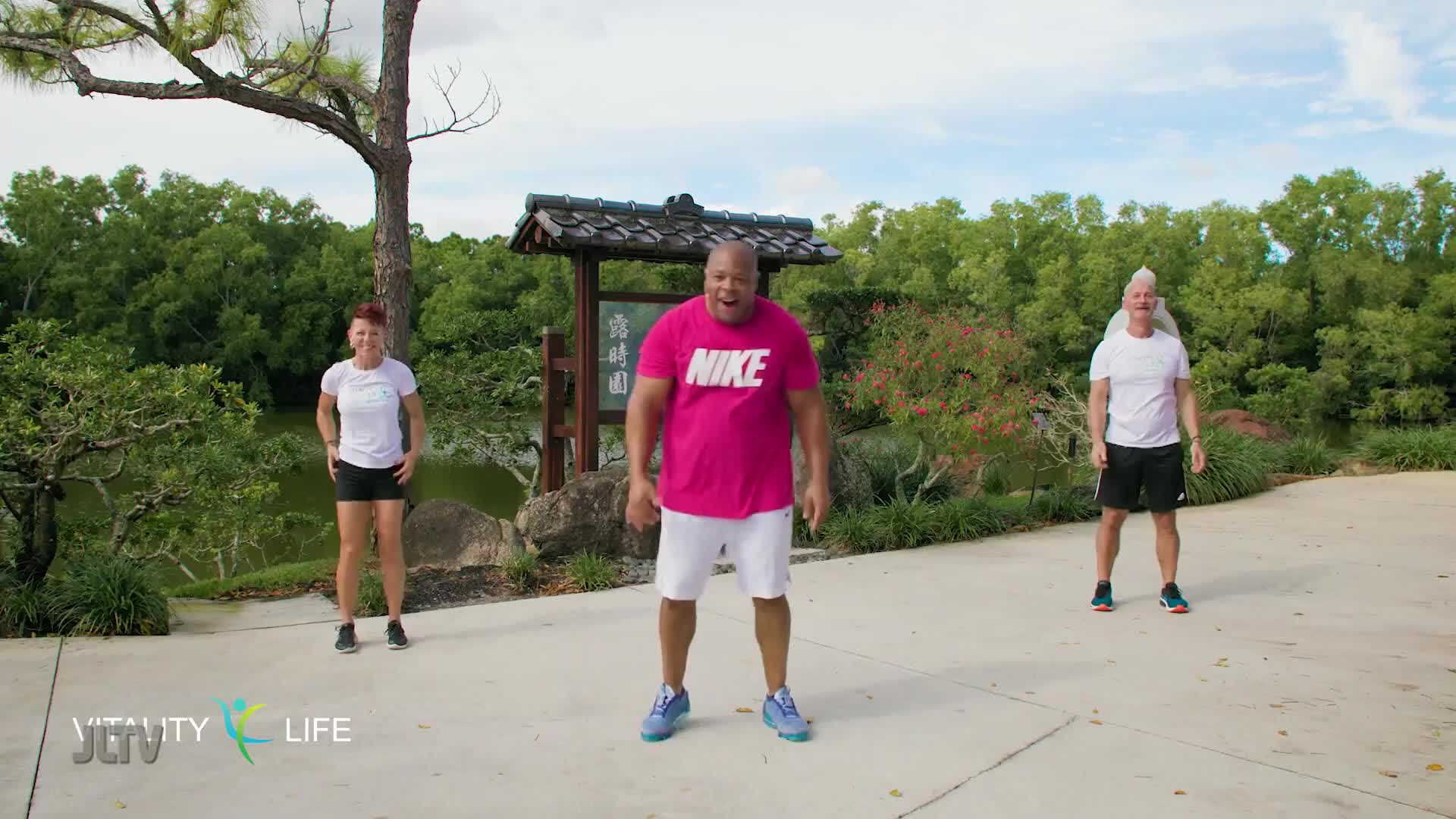 Three people stand in a park, ready to exercise. A woman in a white shirt and black shorts is on the left, a man in a bright pink Nike shirt and white shorts is in the center, and a man with white spiked hair in a white shirt and black shorts is on the right.