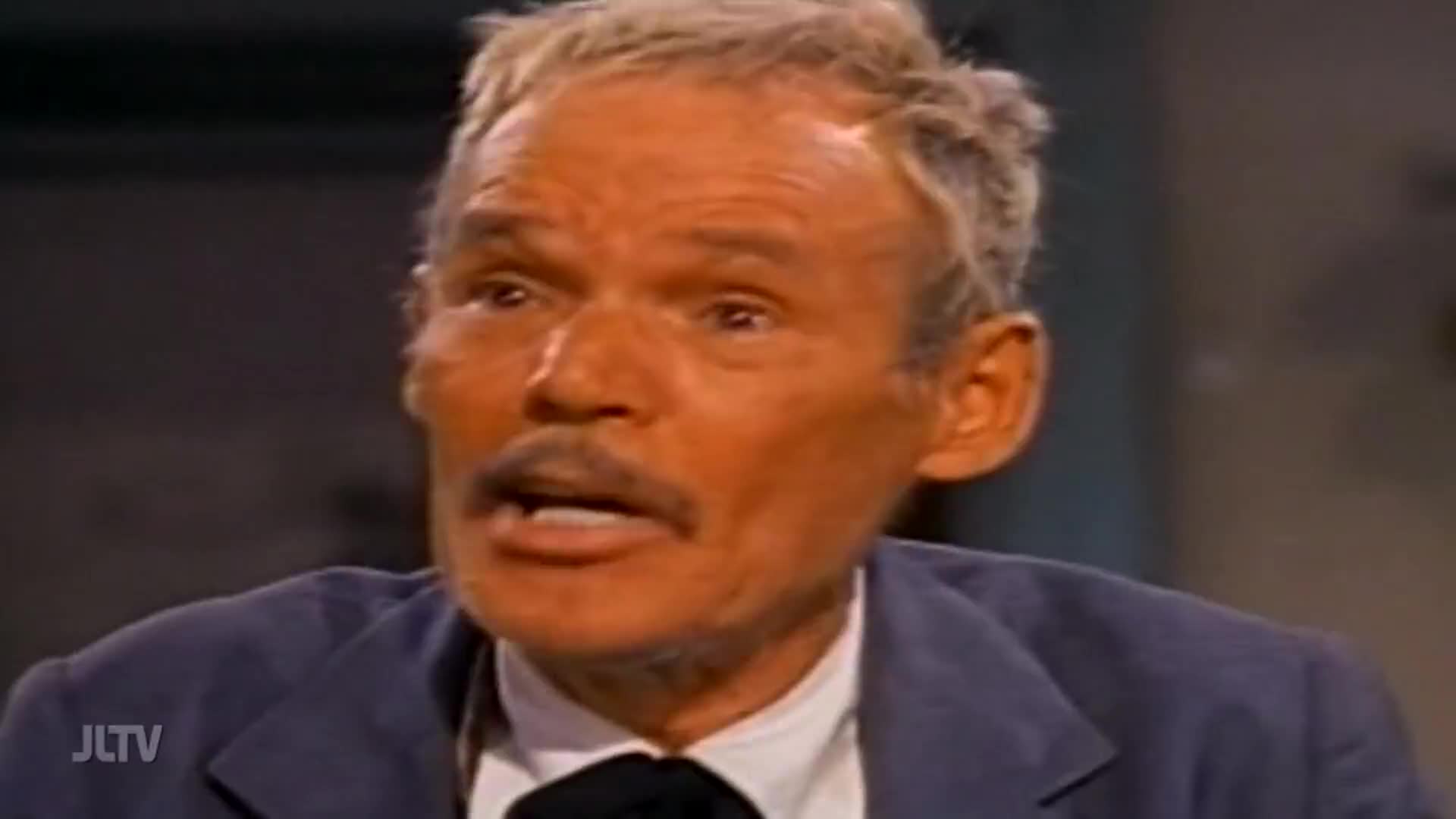 A man with a mustache, wearing a blue jacket over a white shirt and black tie, speaks directly to the camera. The JLTV logo appears in the lower left corner.
