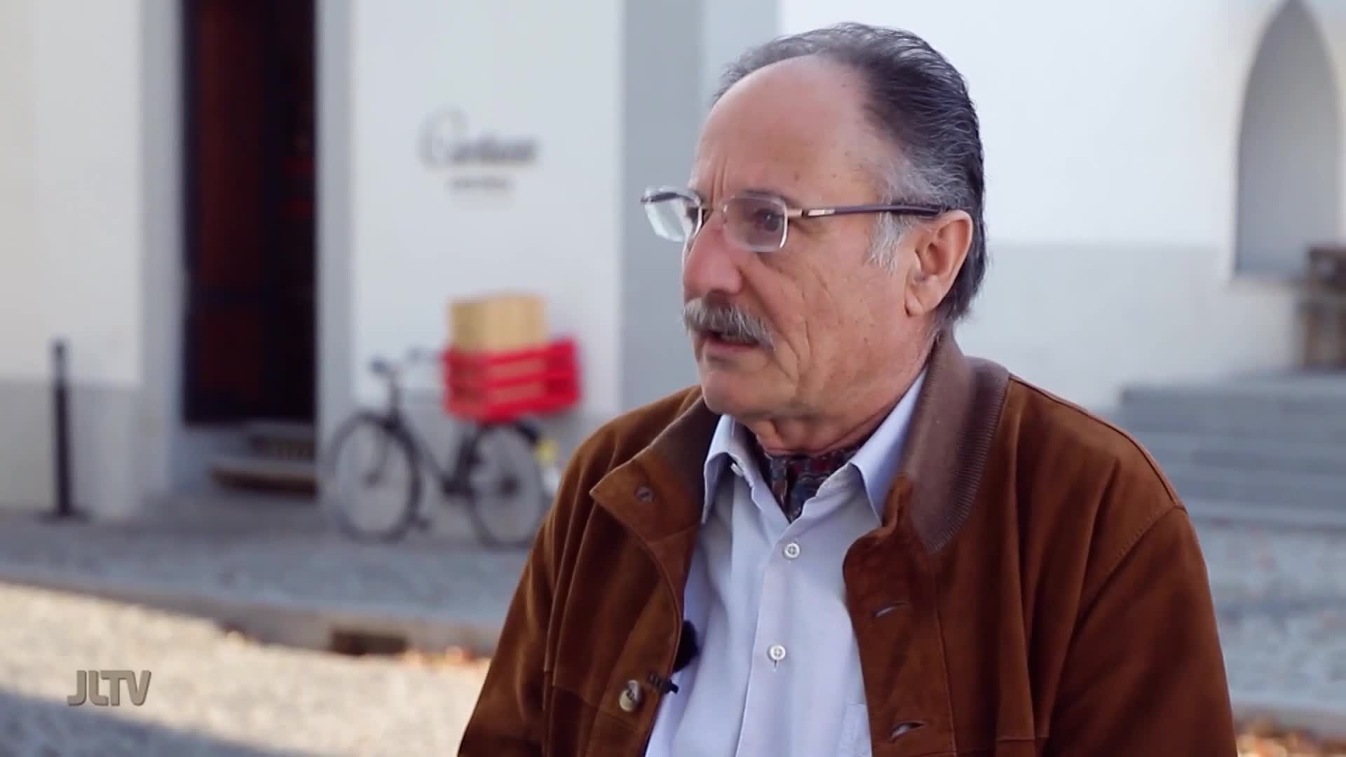 A man with glasses and a mustache speaks, his brown jacket open over a light blue shirt. Behind him, a bicycle with a red basket and a box sits near a building entrance.