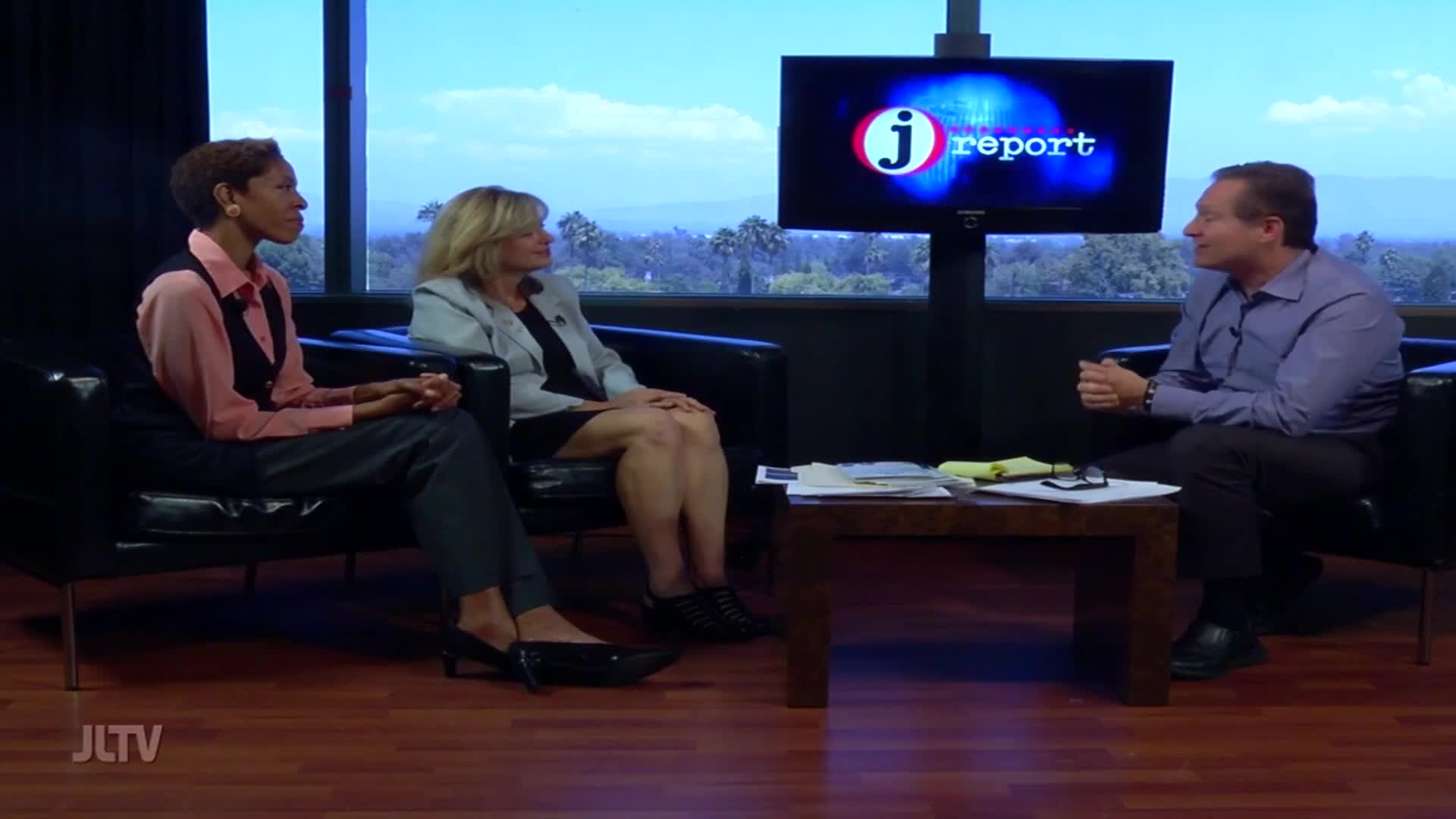 Three people are seated in a studio, facing each other for a discussion. A screen behind them displays the JLTV logo.