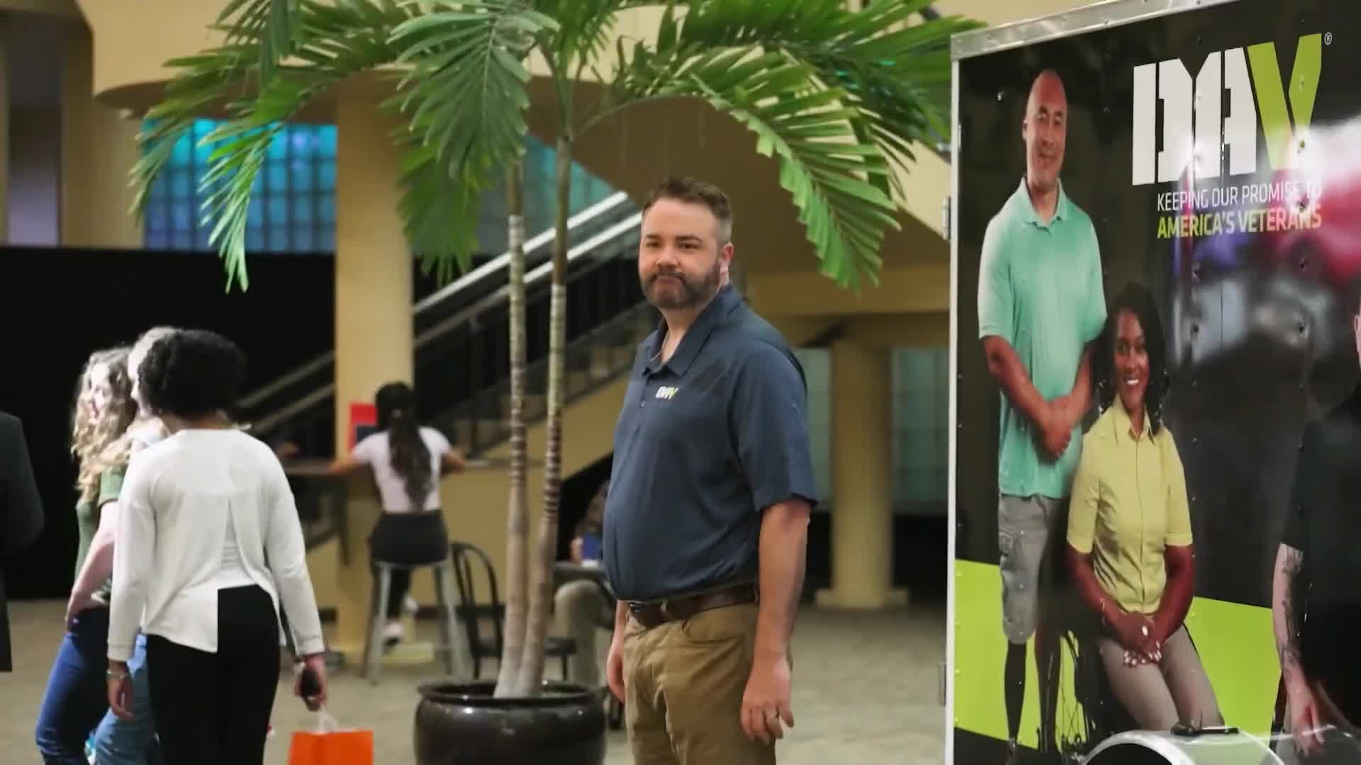 A man in a blue polo shirt stands near a large poster for DAV, an organization supporting America's veterans. Behind him, people mill about in what looks like a community center.