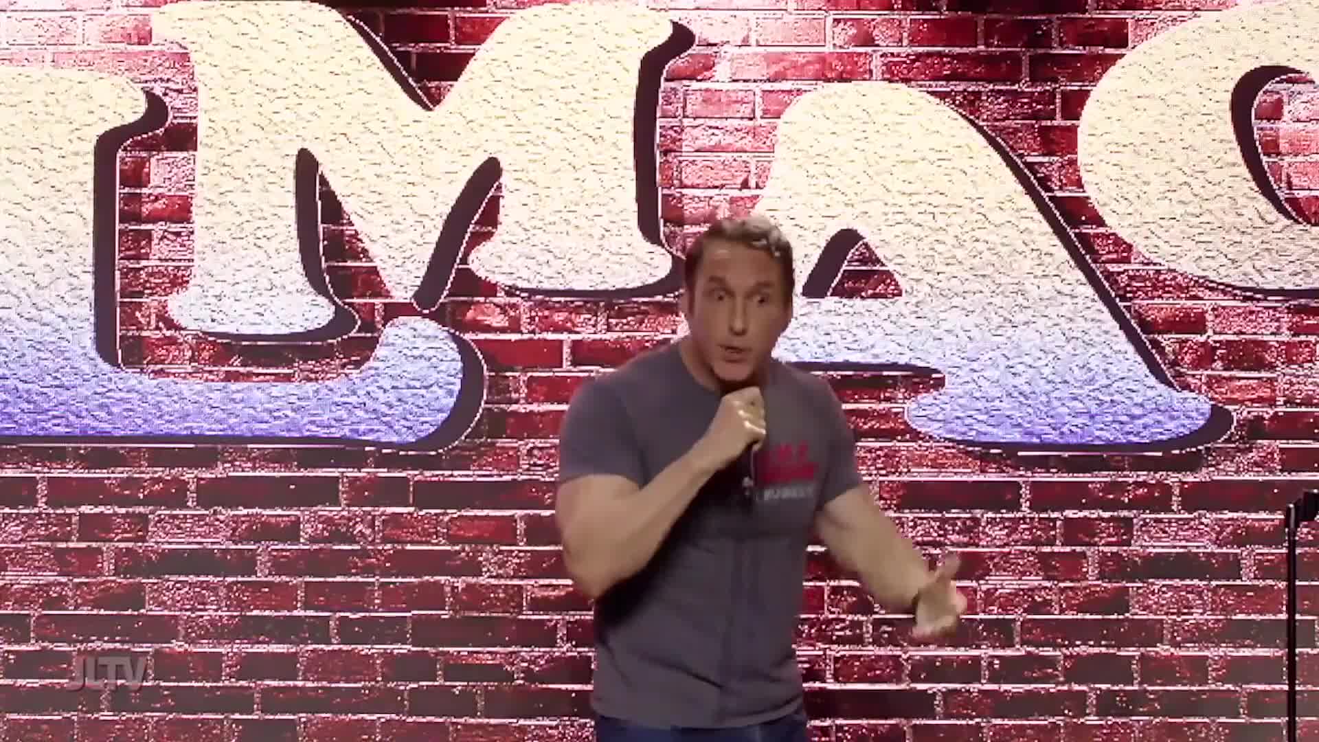 A man stands on a stage, holding a microphone and gesturing with his free hand. Behind him, large, stylized letters spell out "MAC."