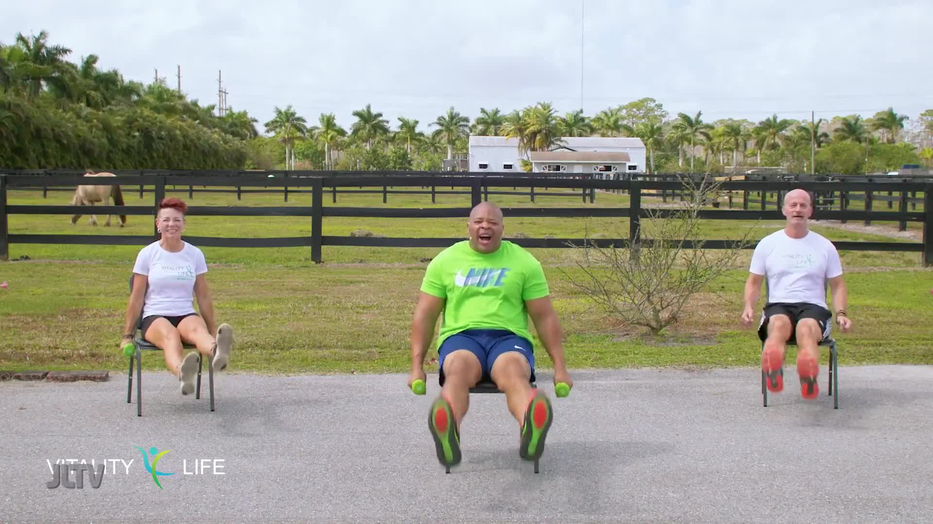 Three people are exercising on chairs outdoors, with a horse grazing in the background. The middle person is holding small weights and appears to be mid-motion, while the others are also seated and engaged in an exercise.