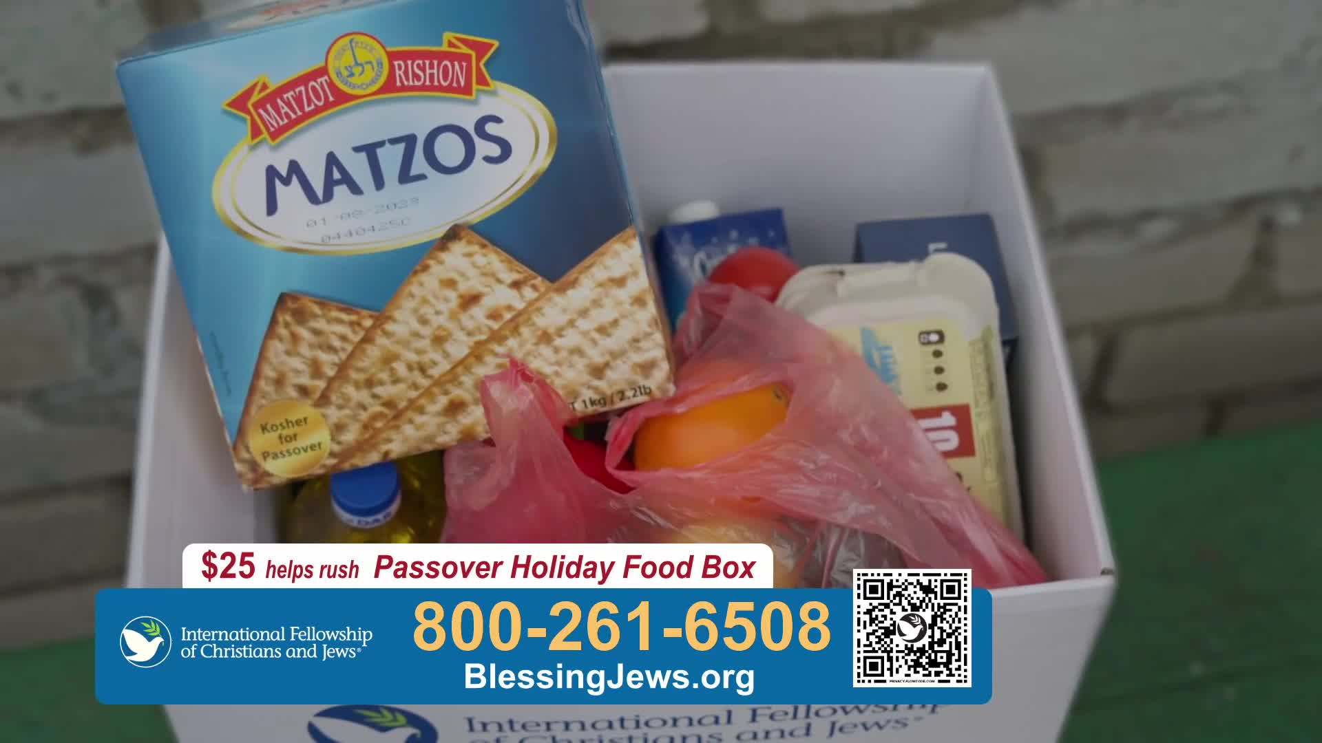 A box overflows with food for Passover, including a large package of matzah. A bright orange sits nestled in a pink plastic bag, peeking out from the other items.