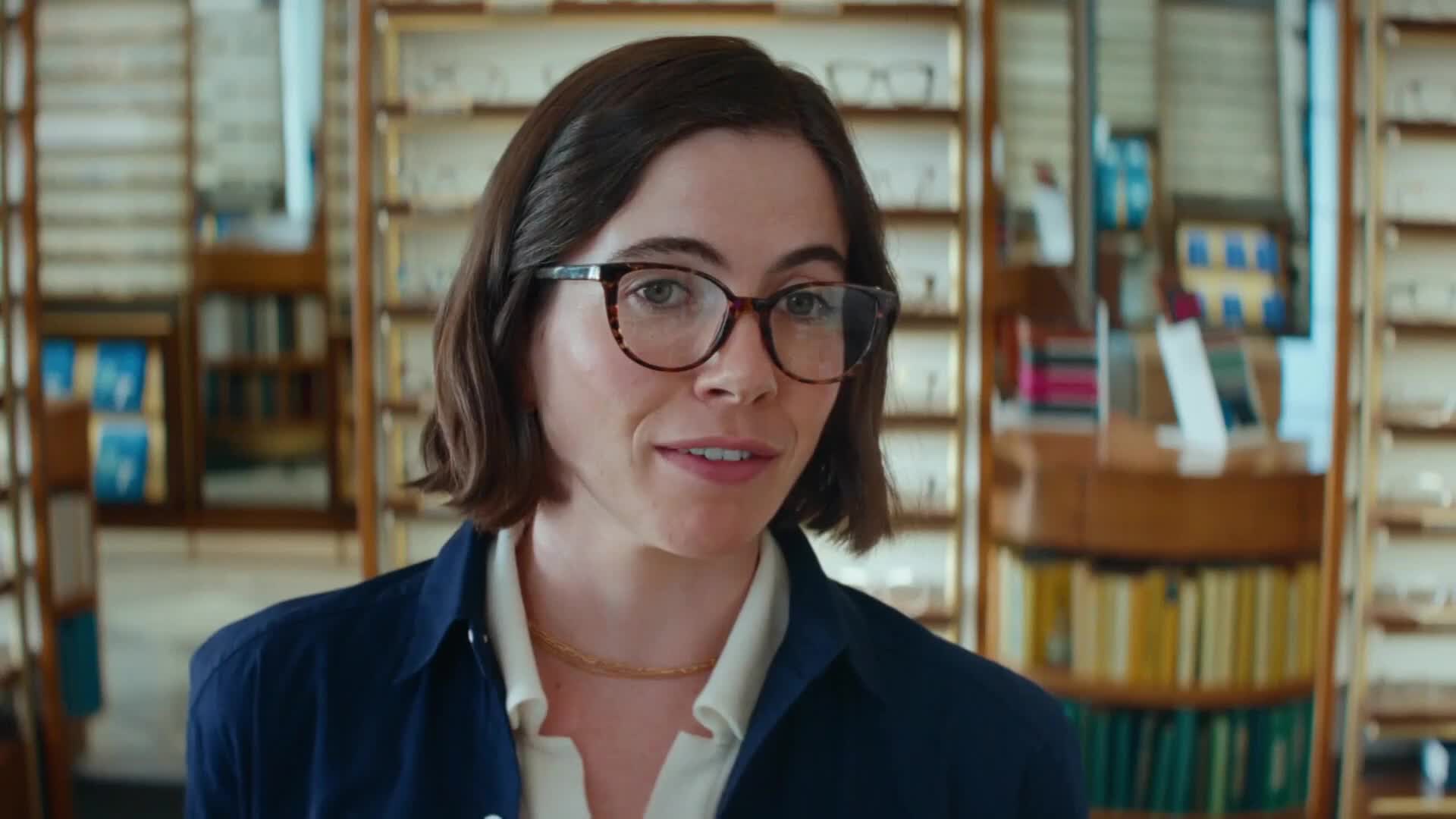 A woman with dark hair and glasses stands in an optical shop. Shelves filled with eyewear surround her.