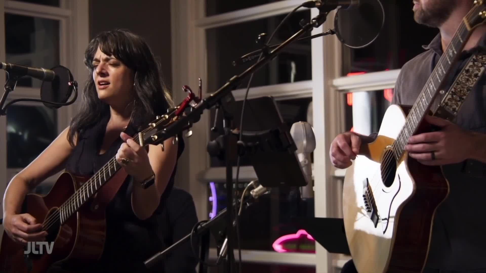A woman sings and strums her acoustic guitar, her eyes closed in concentration. Beside her, a man plays his own guitar, his fingers moving deftly over the fretboard.