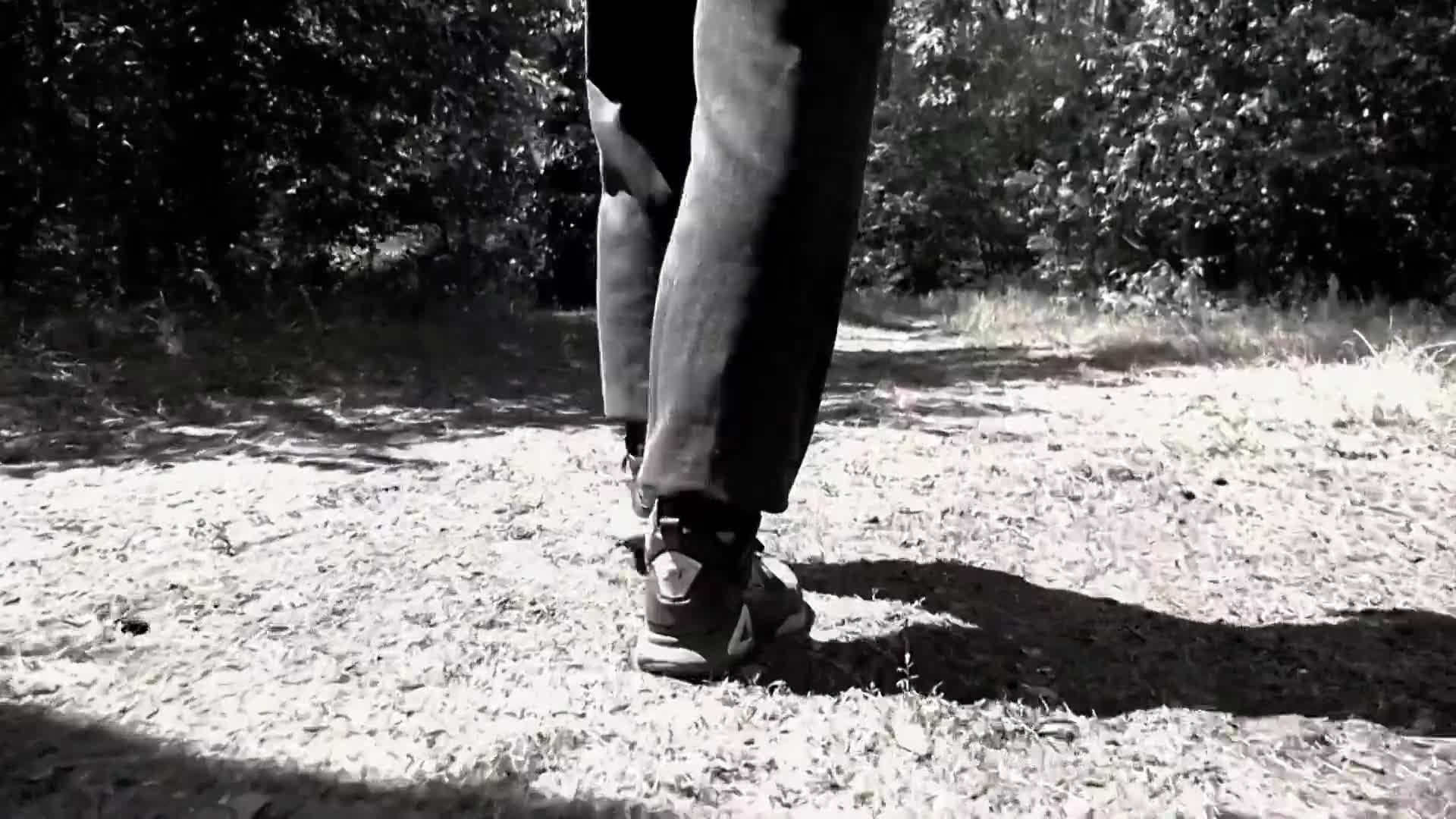 A person walks down a dirt path, their dark pants and sneakers crunching on the gravel. Sunlight filters through the trees, casting dappled shadows on the ground.