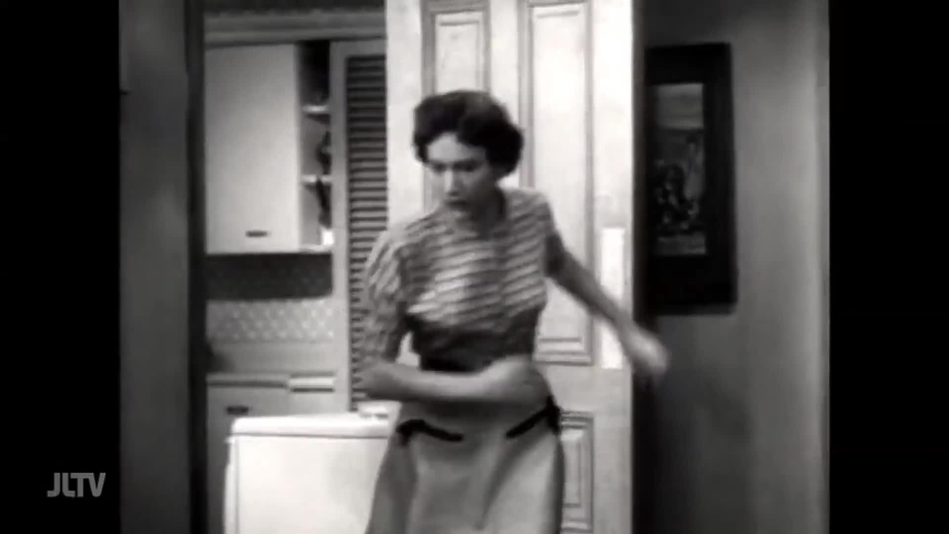 A woman in a striped shirt and apron turns quickly, her arm extended. She's in a kitchen, with cabinets and a refrigerator visible behind her.