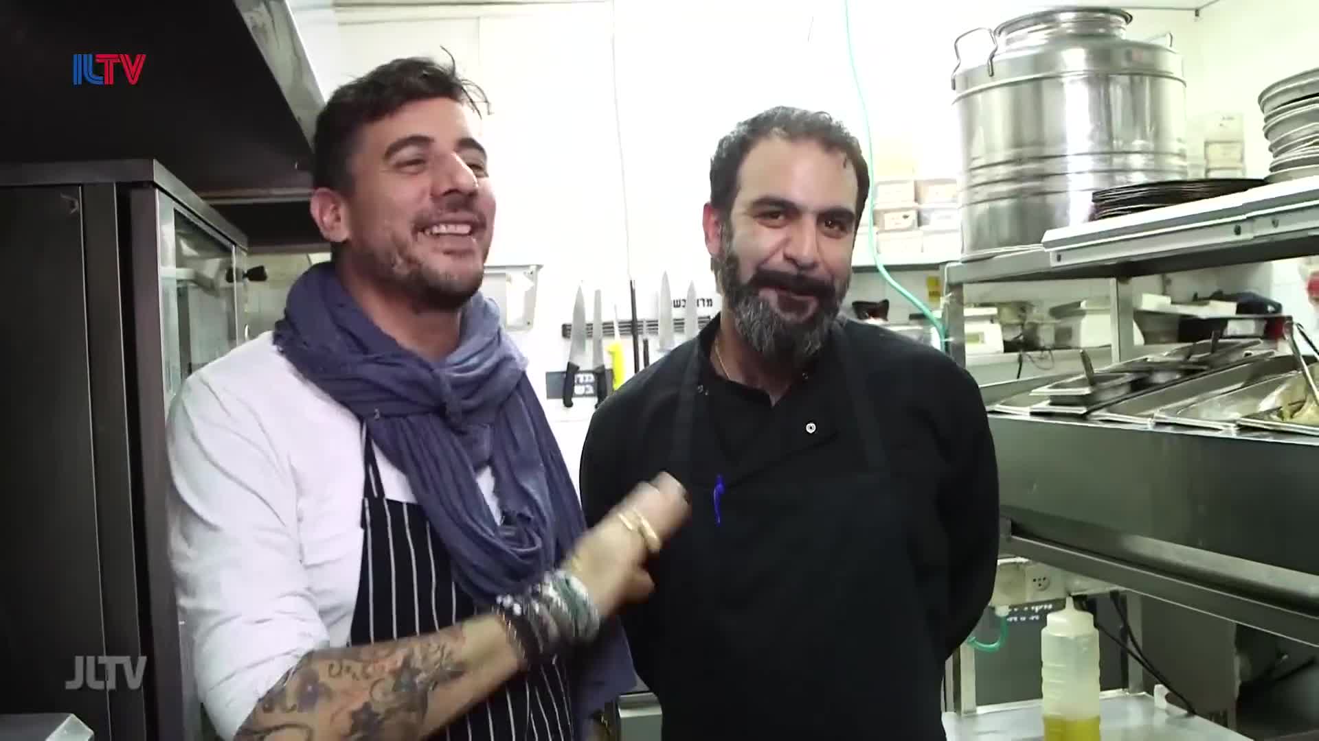 Two men stand in a busy kitchen, one with a blue scarf and striped apron, the other in a black chef's coat. They both laugh, their faces illuminated by the bright lights of the commercial kitchen.