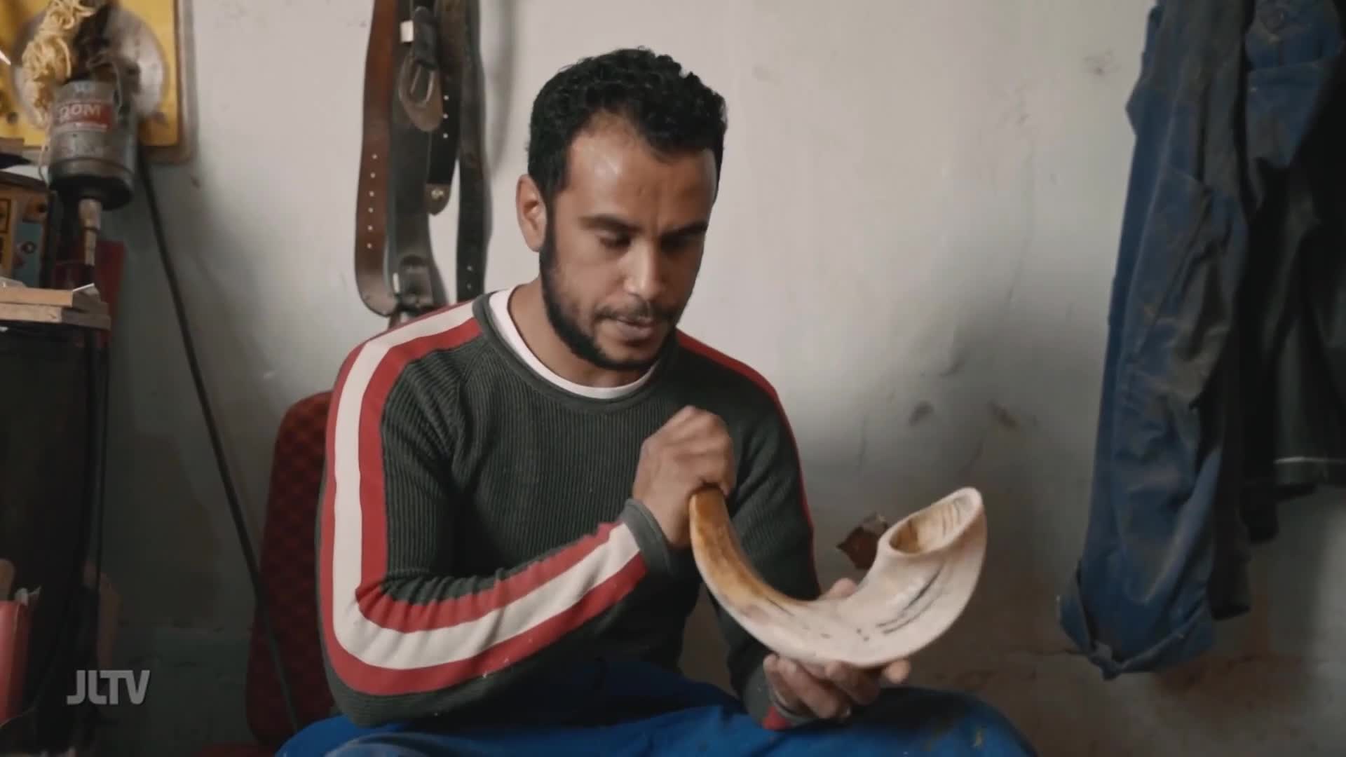 A man carefully holds a shofar, its curved horn gleaming. He is examining the craftsmanship of the instrument, likely preparing it for use in Jewish observances.