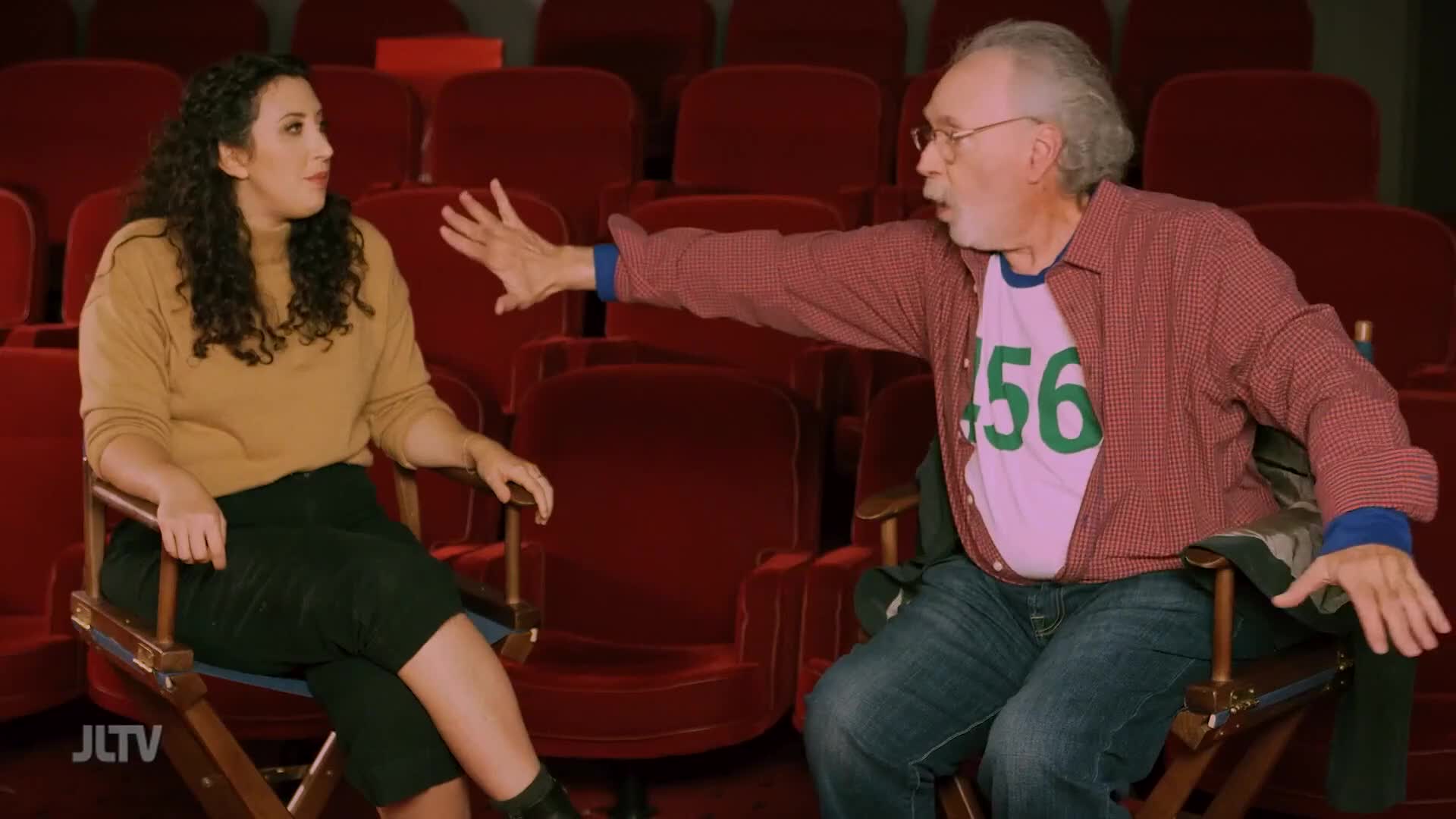A man gestures with his right hand, his arm extended as he speaks. He wears a pink t-shirt with green numbers on it, visible beneath an open red plaid shirt. Beside him, a woman with curly dark hair sits, listening intently.