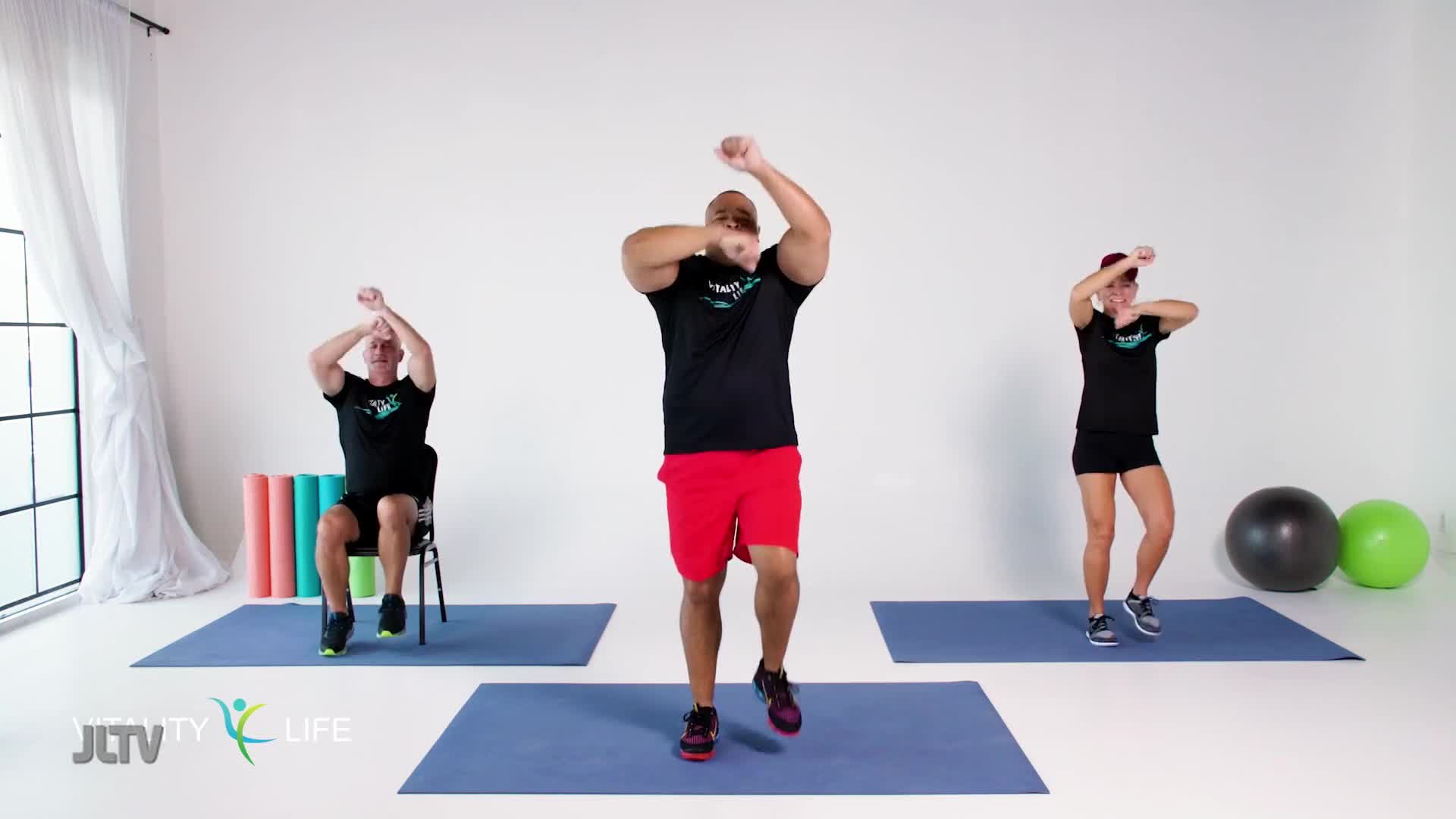 Three people are exercising on blue mats in front of a white wall. The man in the center is doing a high-knee march, while the woman to his right is doing a similar move with bent knees. The man on the left sits in a chair, lifting his knees and pumping his arms.