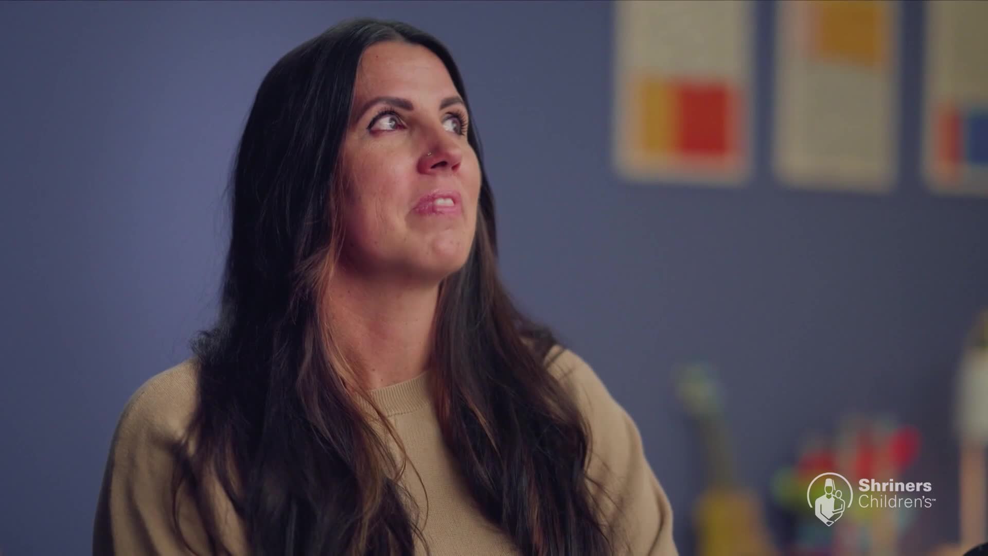 A woman with long, dark hair looks upward with a hopeful expression. Behind her, abstract art hangs on a blue wall, and a Shriners Children's logo is visible.