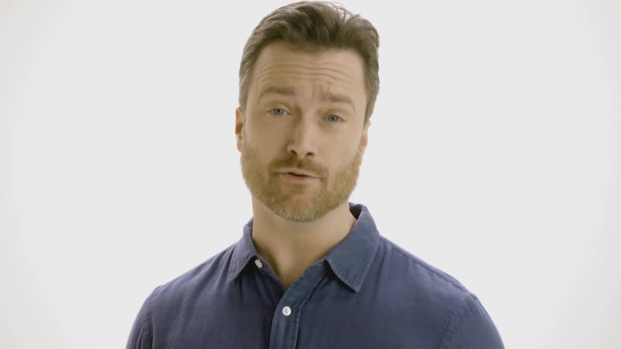 A man in a blue shirt is speaking, his mouth moving as he addresses the camera. He's standing against a plain white backdrop.
