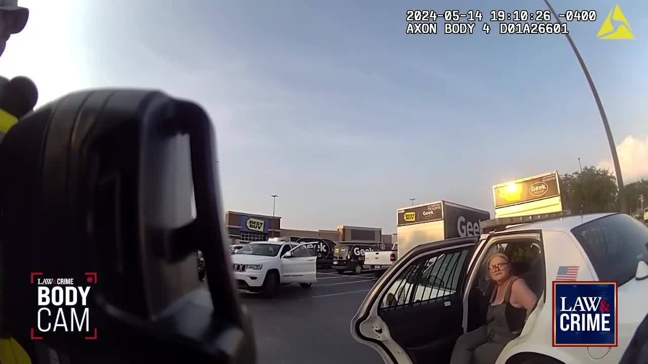 A woman sits in the open back seat of a police car, looking at the camera. The scene unfolds in a parking lot, likely in the United States, as indicated by the "Law & Crime" body cam footage.
