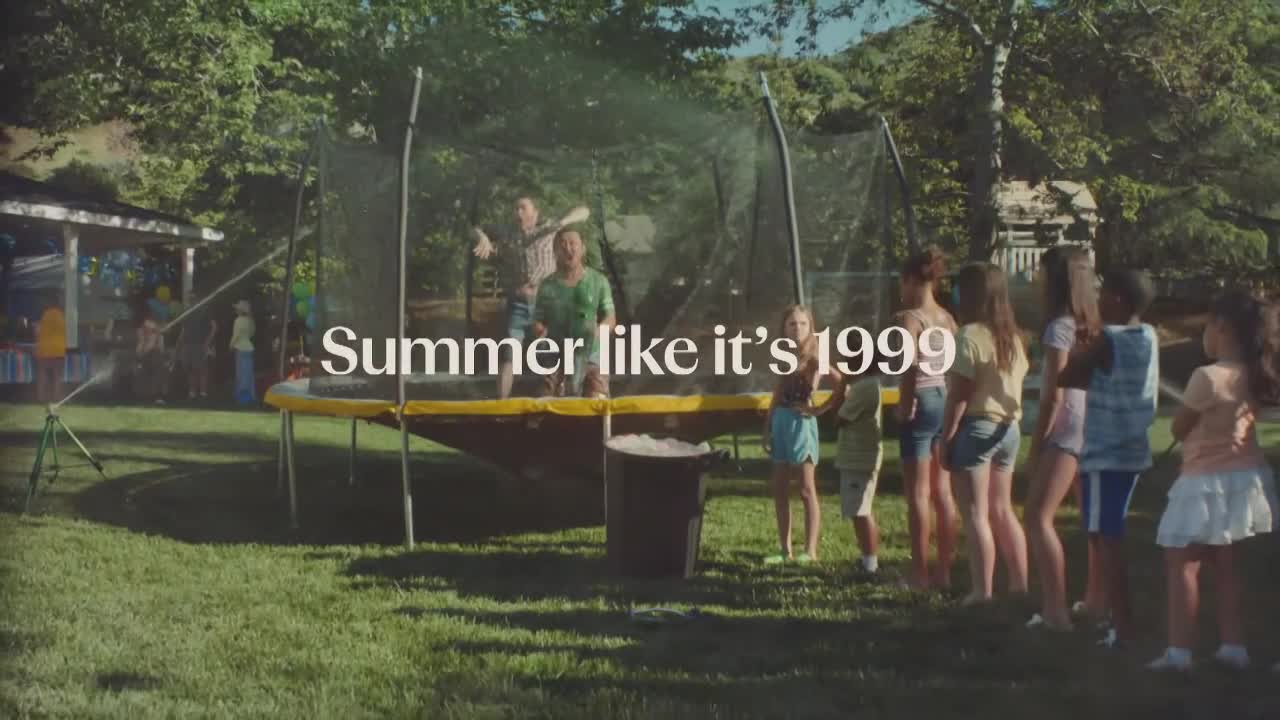 Two guys are jumping on a trampoline, getting sprayed by a sprinkler. A group of kids watch from the grass, while the words "Summer like it's 1999" are superimposed over the scene.
