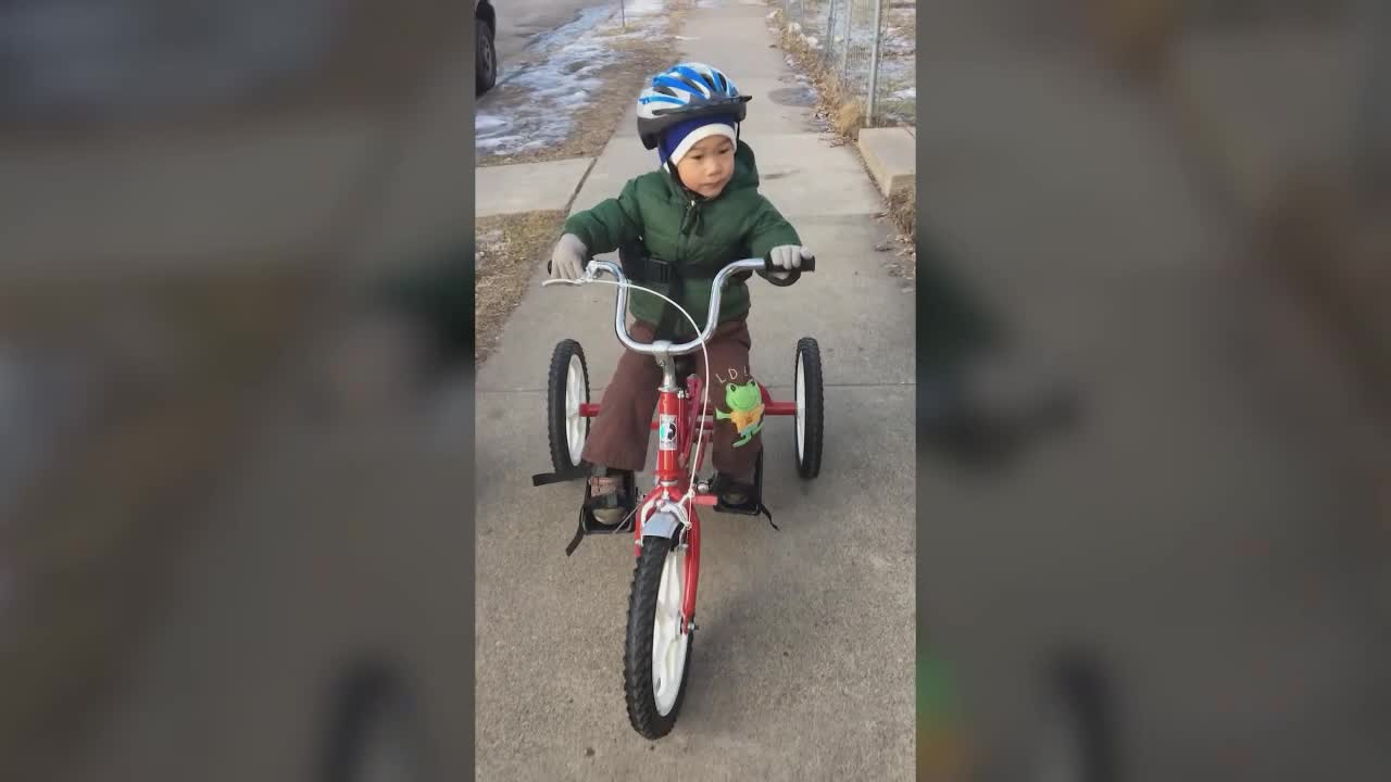 A young child in a blue helmet pedals a red tricycle down a sidewalk. He's wearing a green jacket and brown pants, and a small frog toy is clipped to the handlebars.
