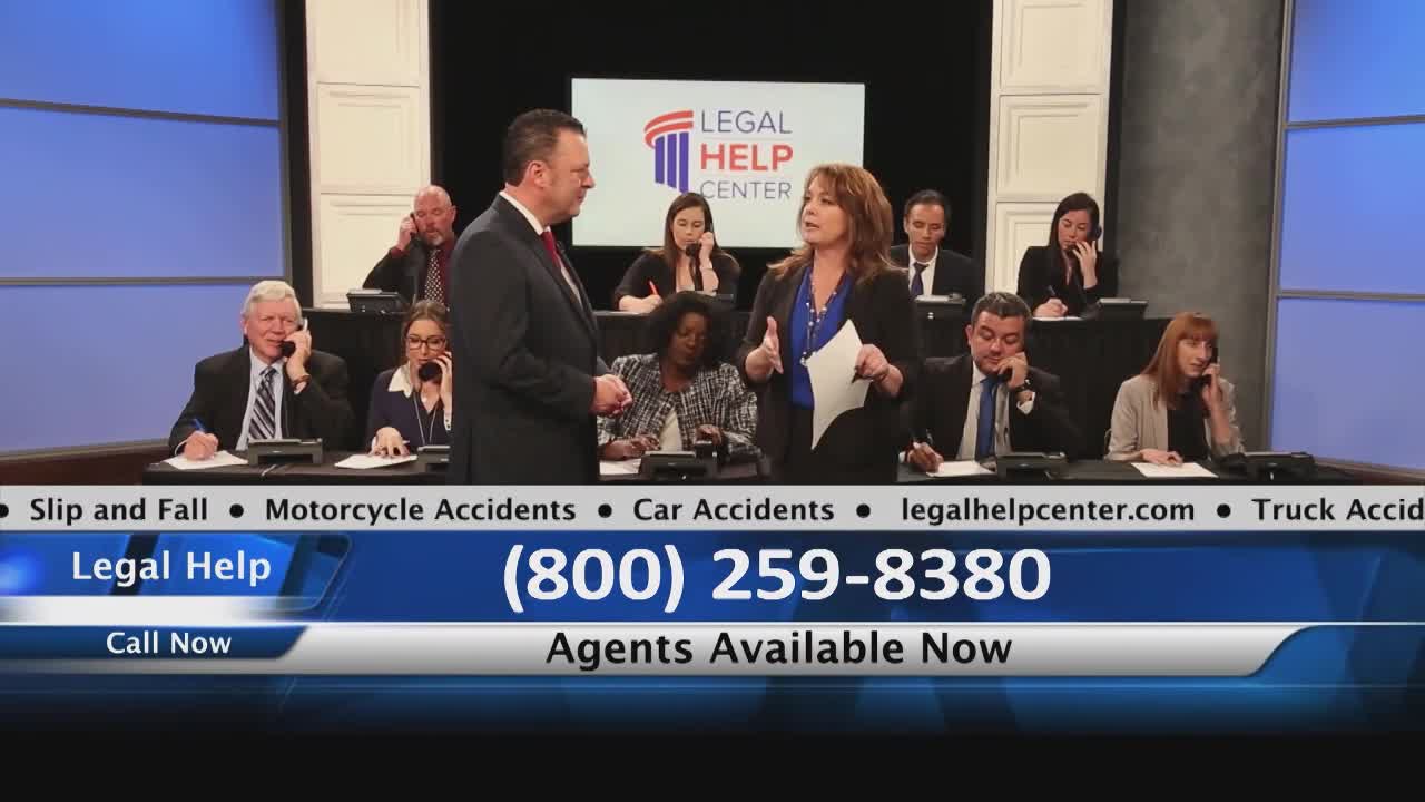 A woman in a blue top gestures while holding a paper, speaking to a man in a suit. Behind them, several people are on the phone at their desks, likely taking calls for a legal help center.

