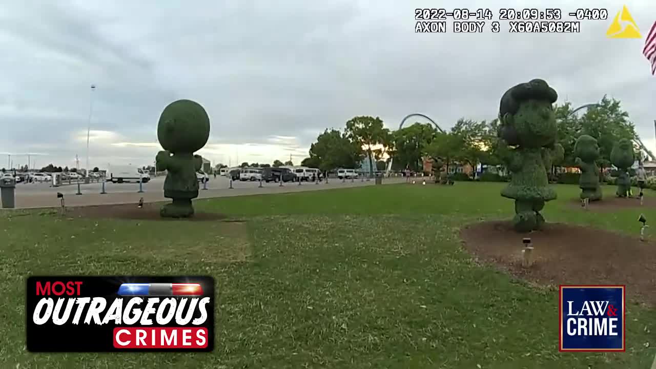 The camera angle shows a grassy area with topiary figures of cartoon characters. Beyond them, a parking lot and roller coaster are visible, suggesting a theme park setting.
