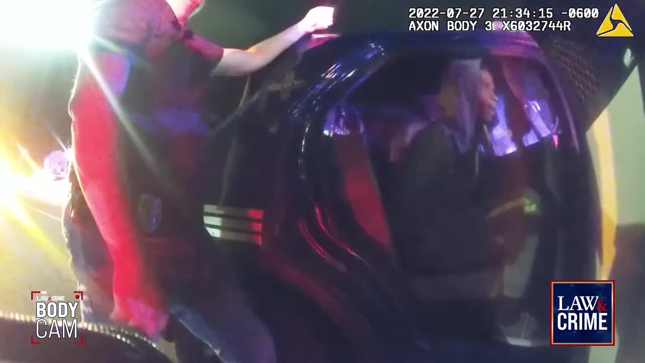 A police officer is leaning into a car, his arm resting on the roof. Inside, a person is visible, illuminated by the flashing lights of the police vehicle.
