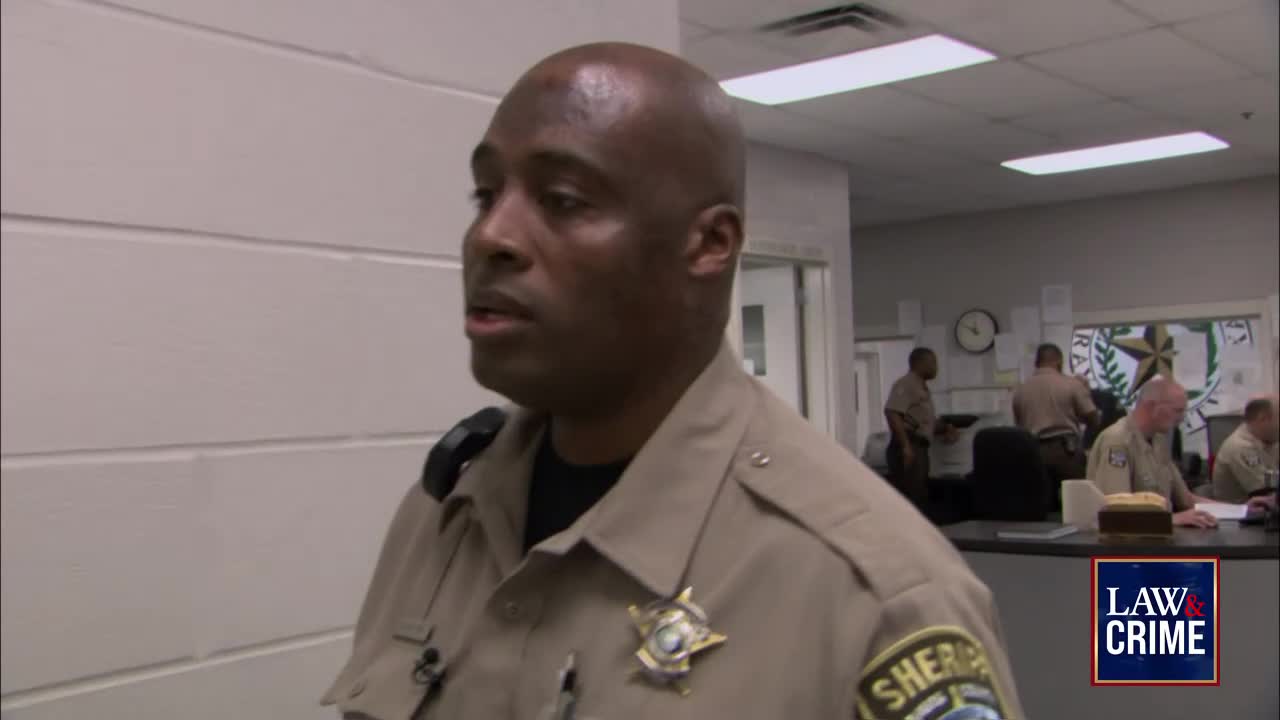 A sheriff in uniform is speaking, his gaze directed slightly to the side. Behind him, other officers are working in a brightly lit office, and the "Law & Crime" logo is visible.
