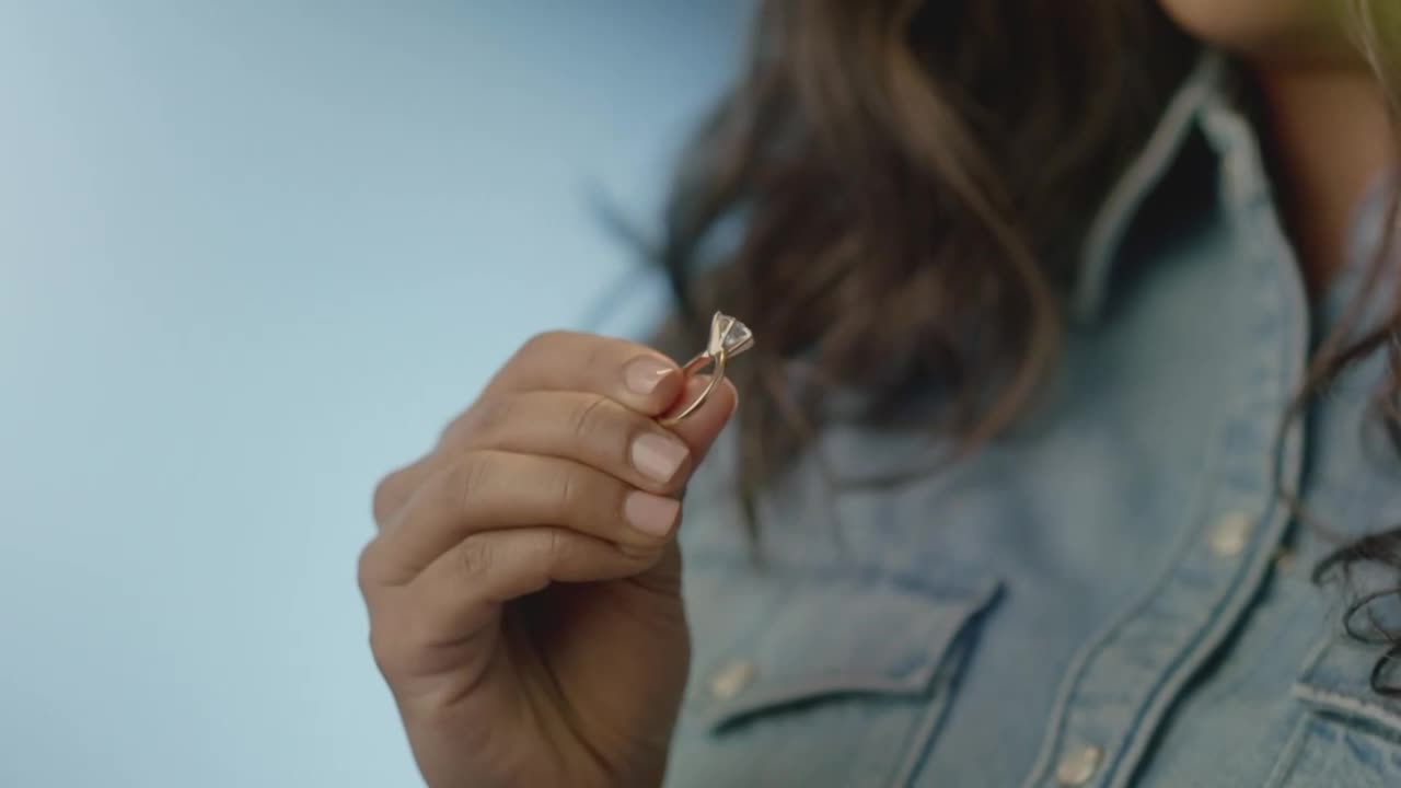 A woman's hand holds up a diamond engagement ring, the light catching the facets. She's wearing a denim shirt, and her dark hair frames her face.
