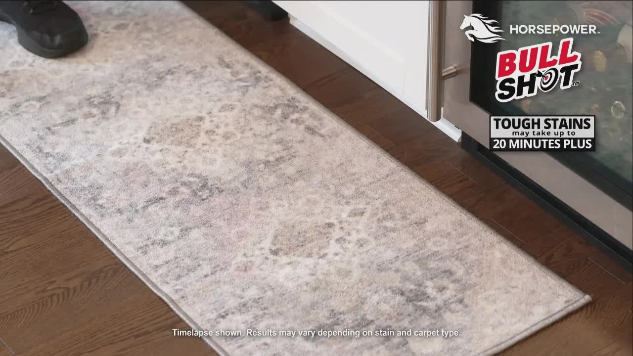 A foot in a black shoe is stepping onto a patterned rug. The rug is placed on a dark wood floor, next to what looks like a refrigerator.
