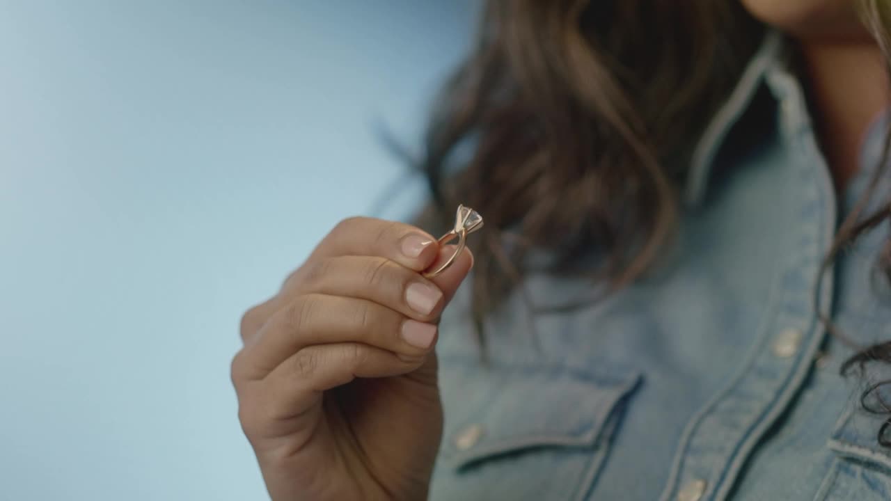 A woman holds up a sparkling diamond ring, the light catching its facets. She's wearing a denim jacket, and the background is a soft blue.

