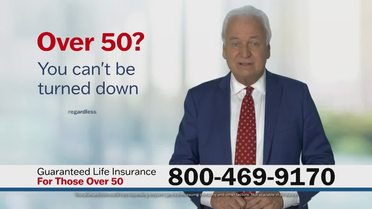 A man in a blue suit and red tie stands in front of a blurred background, speaking directly to the camera. Text on the screen advertises guaranteed life insurance for those over 50.
