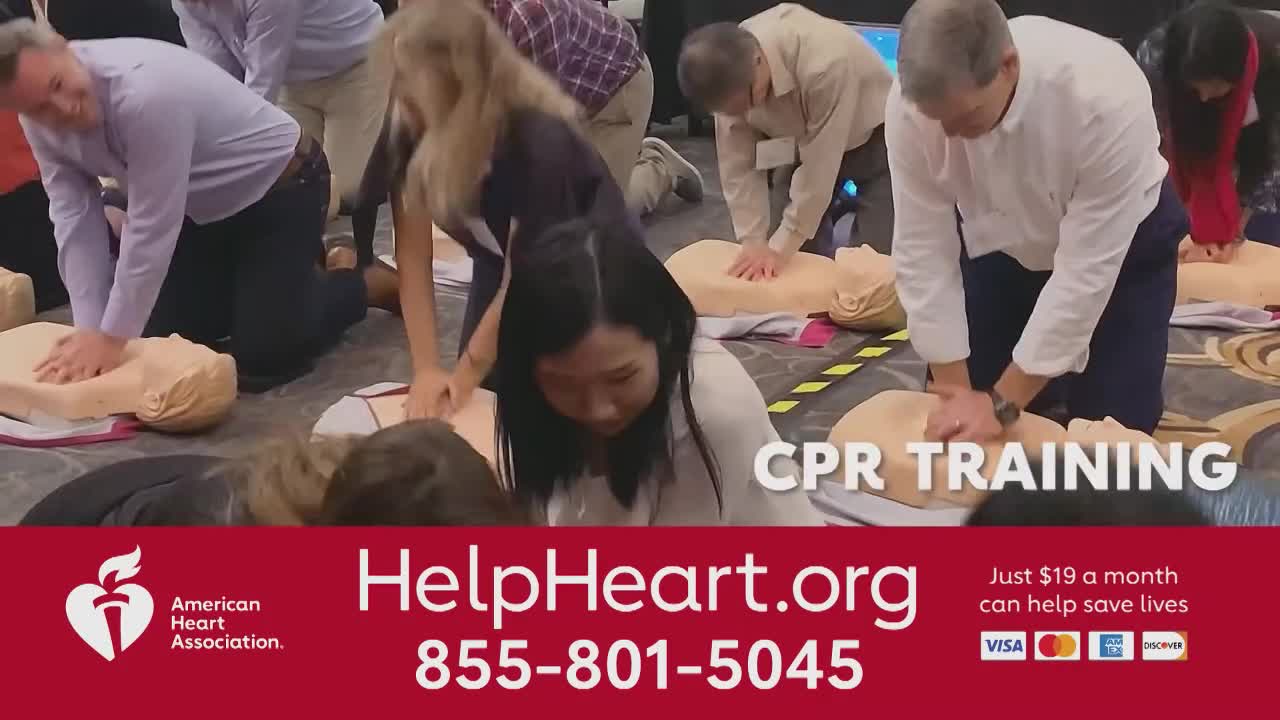 People are kneeling around mannequins, their hands pressing down on the chests. It's a CPR training session, with the American Heart Association logo visible.
