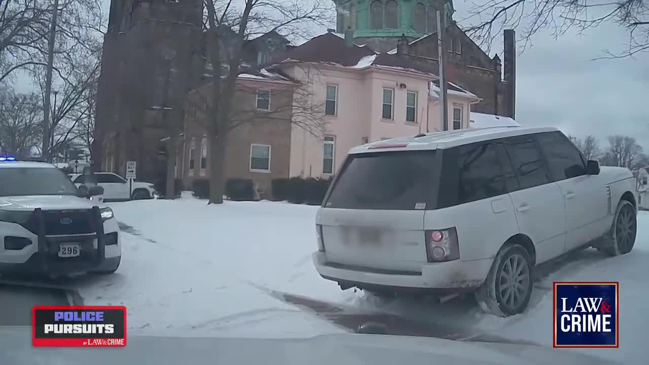A white SUV is angled on a snow-covered road, its rear tires kicking up slush. A police cruiser with flashing lights is parked nearby, and the scene is being covered by Law & Crime.
