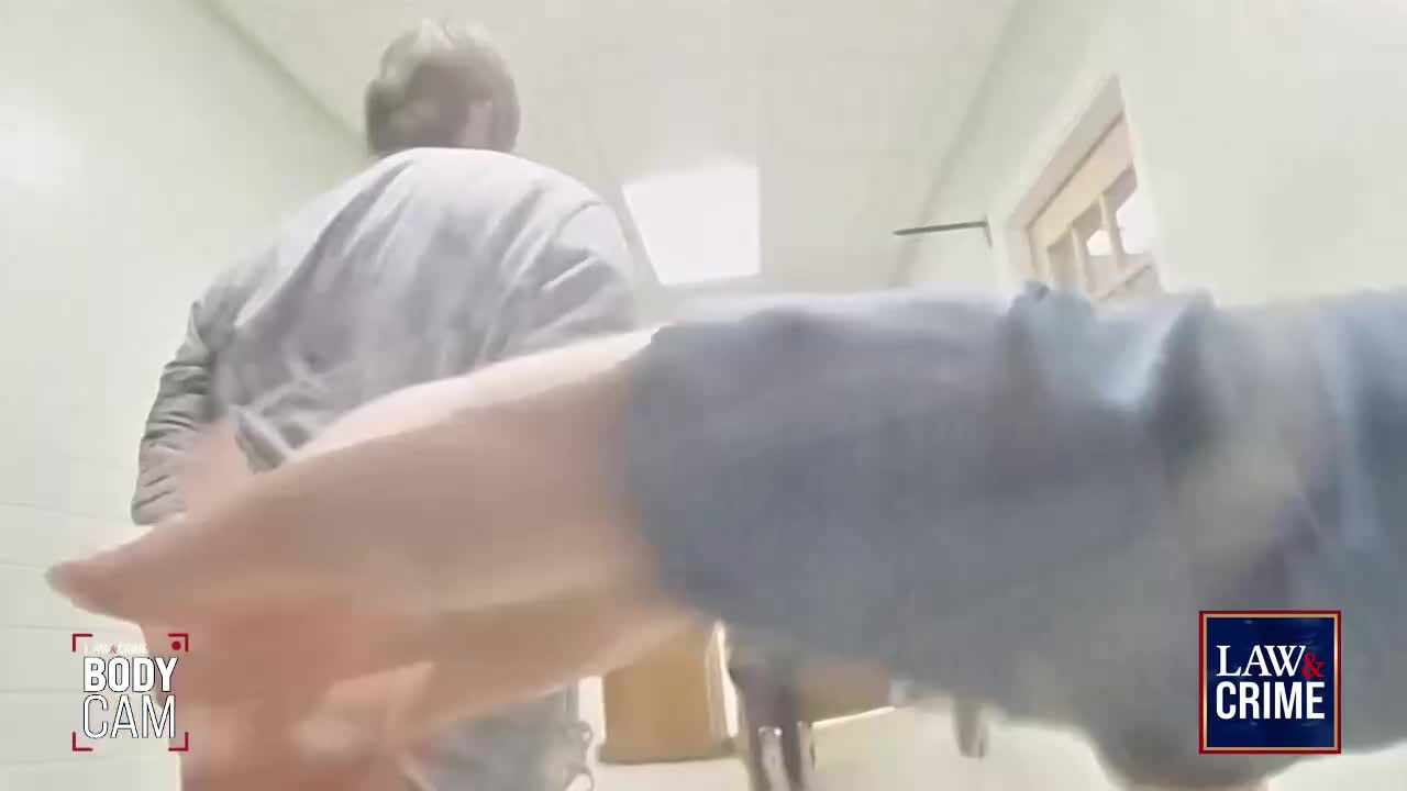 A man in a gray shirt is walking away, his hands cuffed behind his back. The camera, likely a body cam, is pointed forward, and another person's arm is visible in the foreground.
