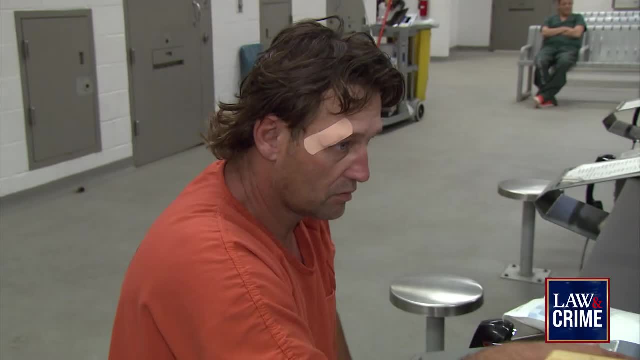 A man in an orange jumpsuit sits at a table, a bandage on his face. Across the room, a man in green scrubs sits on a bench, seemingly waiting.
