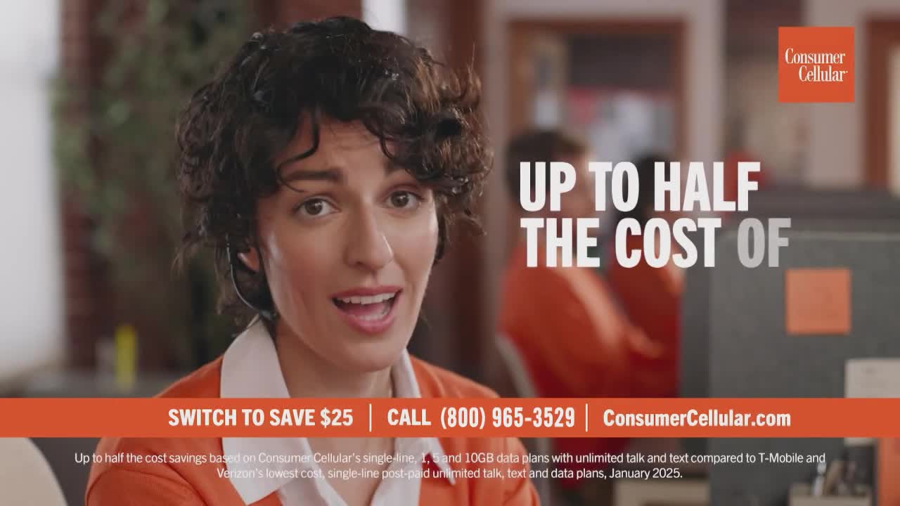 A woman in a headset smiles, speaking directly into the camera. Behind her, another person in an orange shirt is blurred.

