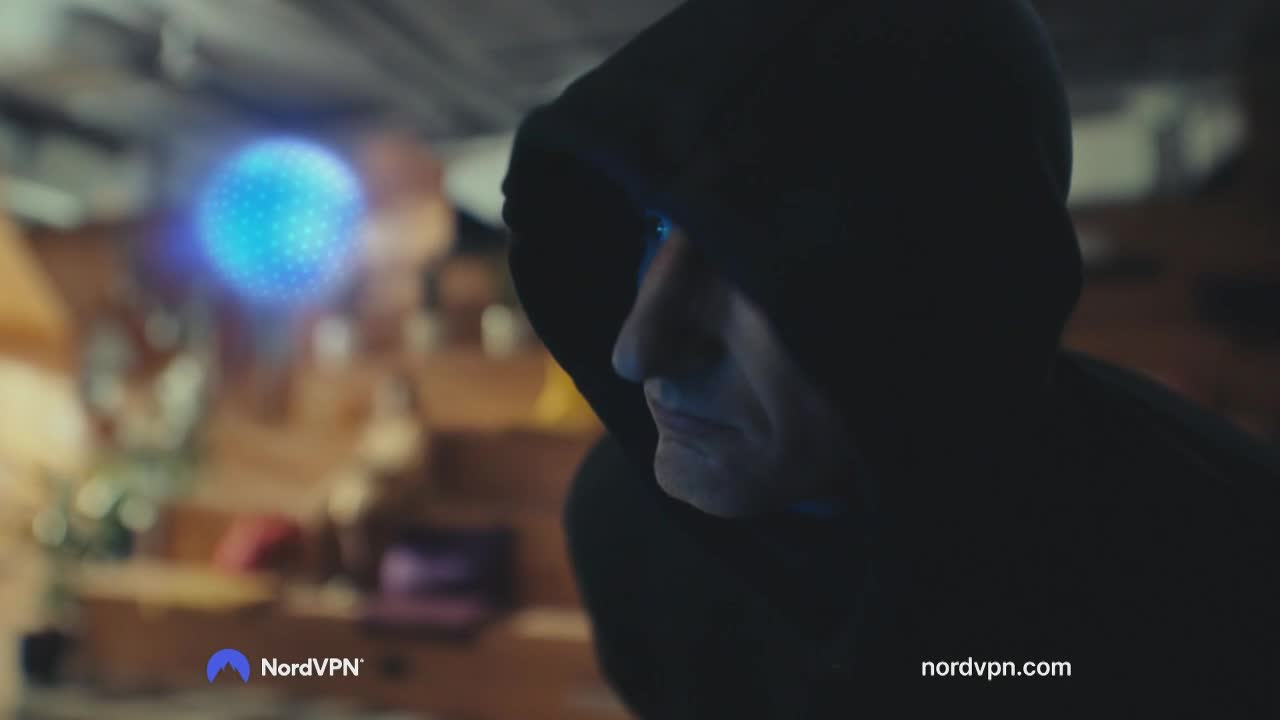 A hooded figure is in profile, their face lit with a cool blue glow. Behind them, a bright, spherical light pulsates, suggesting a digital or technological environment.
