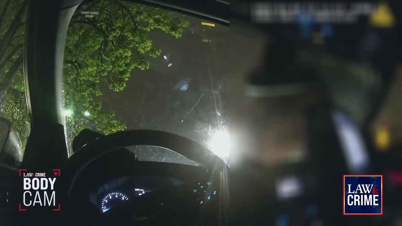 Outside the windshield, a bright light shines through the rain, illuminating the tree branches. The body cam footage, branded by Law & Crime, is from inside a car, showing the steering wheel and dashboard.
