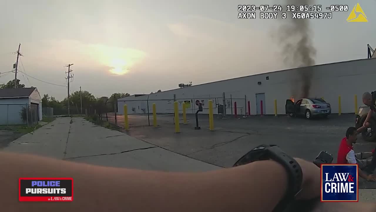 A car is engulfed in flames, thick black smoke billowing upwards. Officers and civilians are gathered nearby, likely responding to the emergency.
