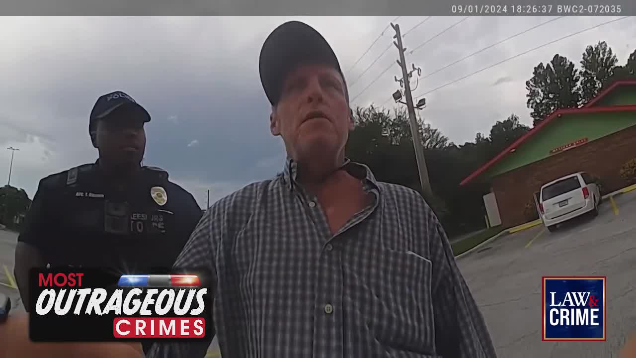 A man in a plaid shirt stands in front of a police officer, both facing the camera. The scene appears to be unfolding outside a building, likely in the United States, as indicated by the Law & Crime logo.
